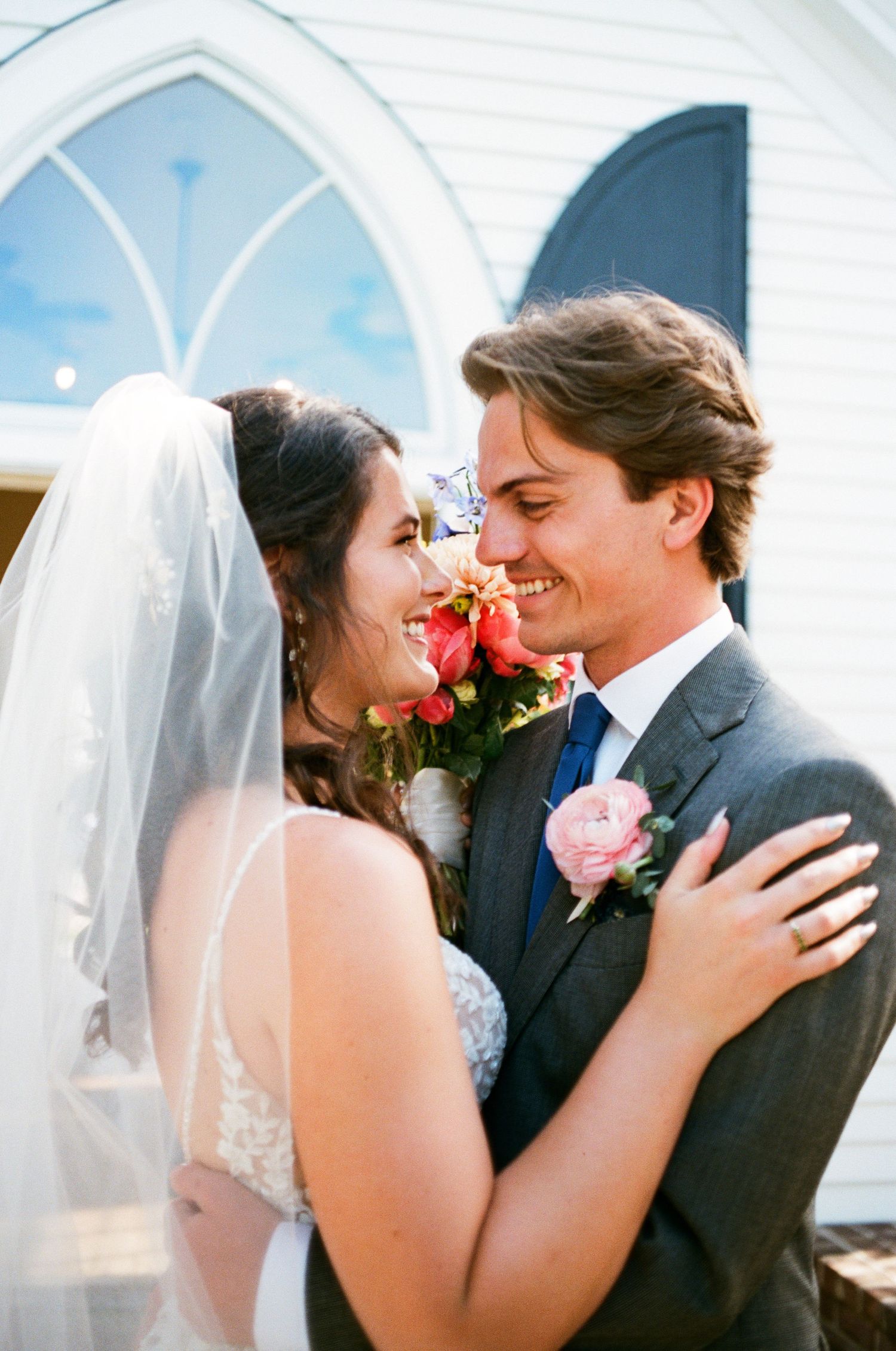 A happy bride and groom share an intimate moment outside a chapel with white arched doorway and pink rose boutonniere.