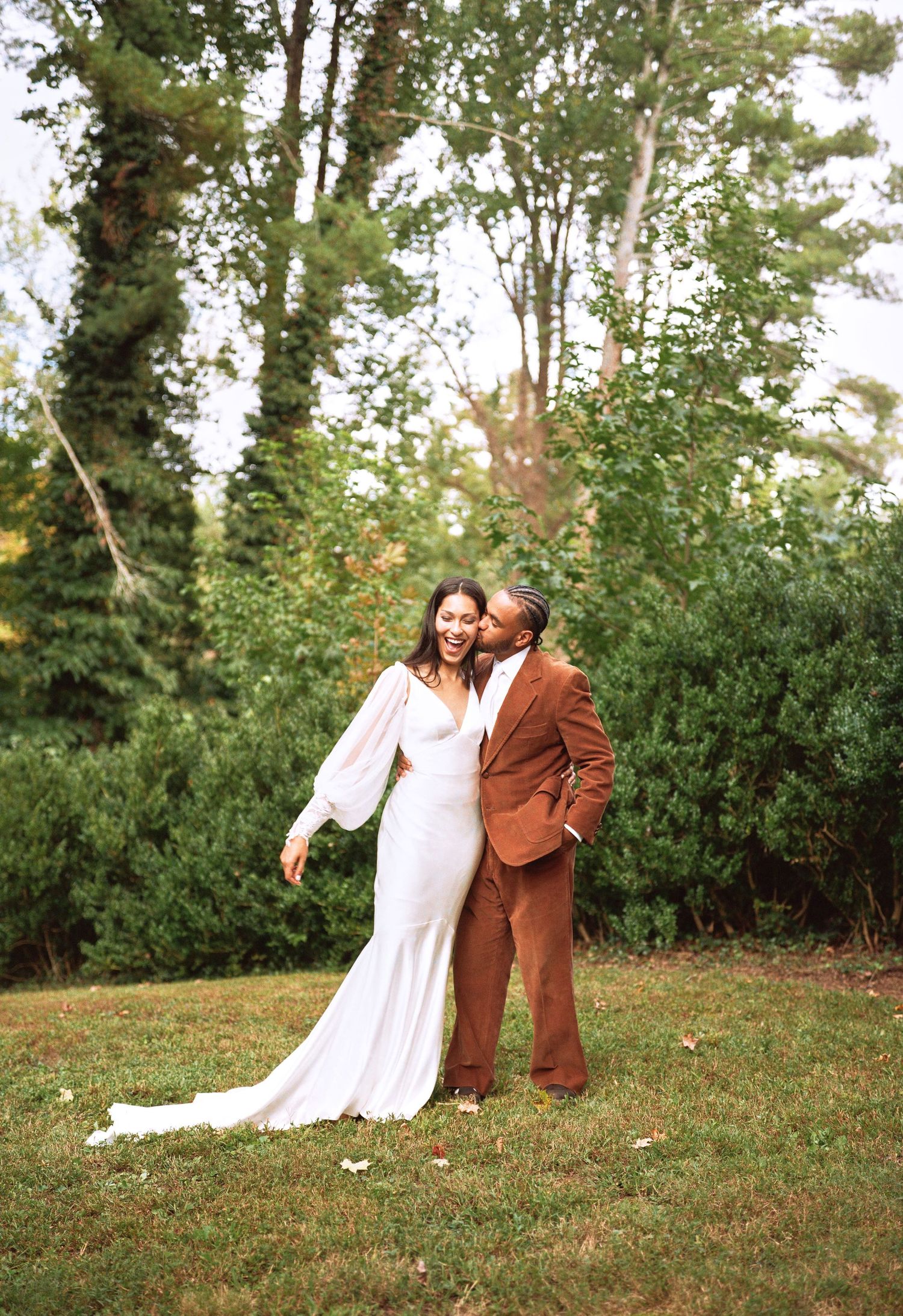 A couple in wedding attire embraces on a grassy lawn surrounded by lush greenery.