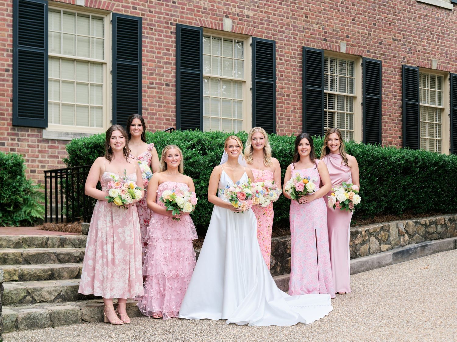 Bridesmaids in pink floral and solid dresses pose with bride in white gown outside brick building.