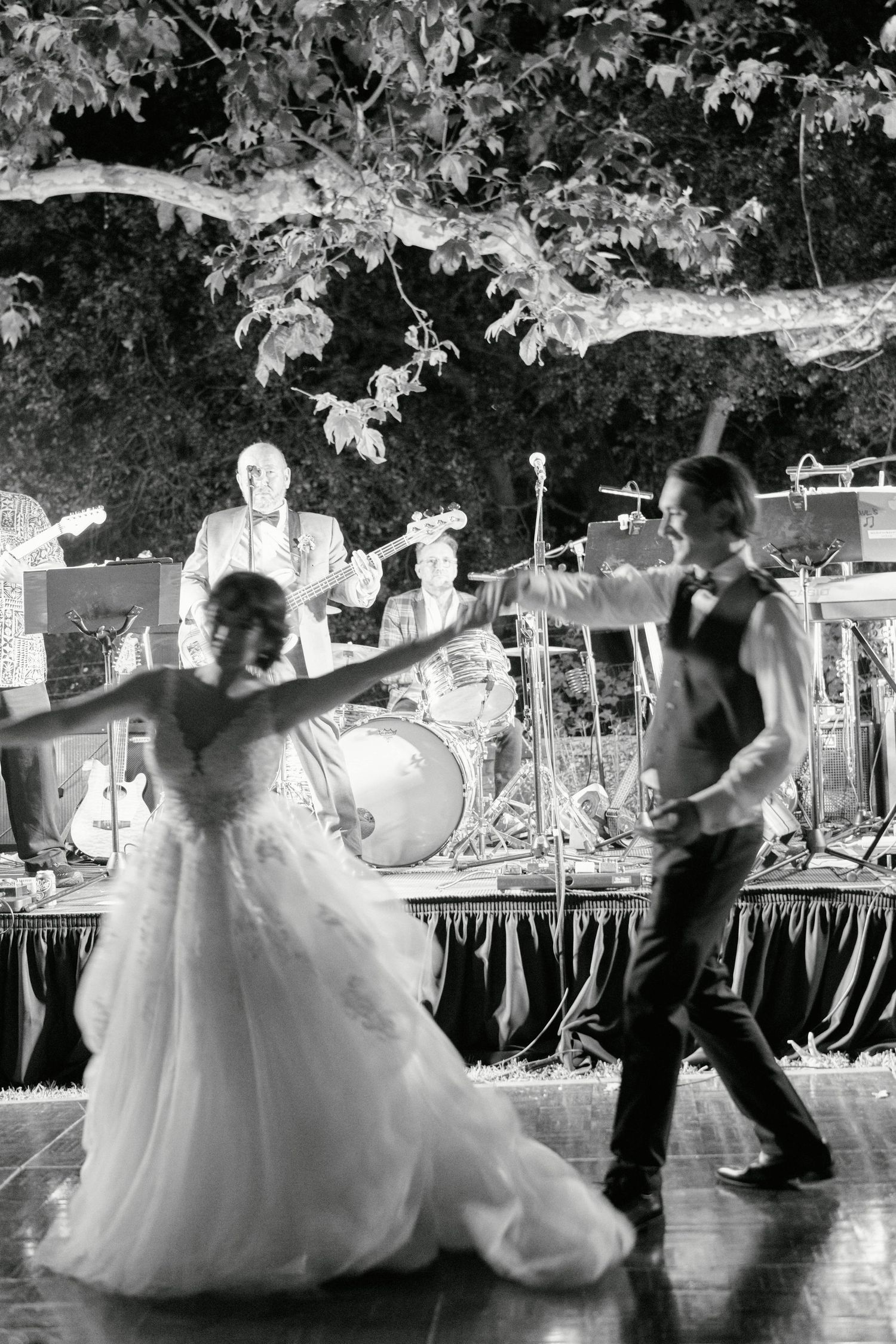 A couple dances joyfully at their outdoor wedding reception with musicians performing in the background.