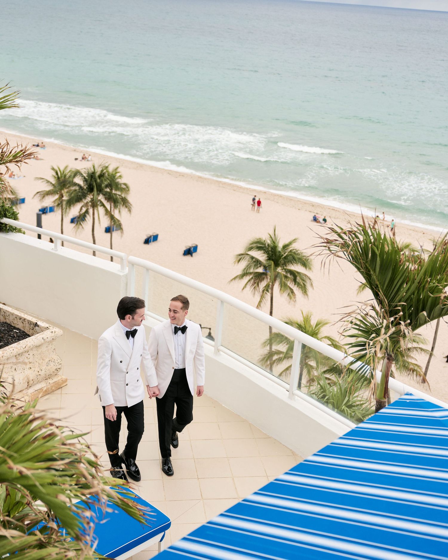 Two people in formal attire walk down a blue-carpeted walkway with palm trees and beach views.