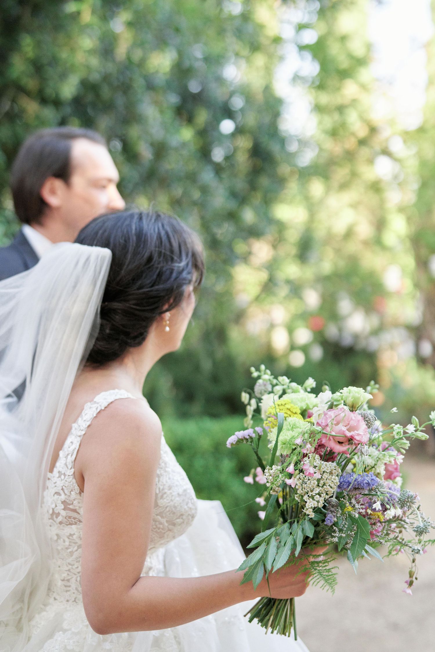 A bride in a lace wedding dress holds a beautiful wildflower bouquet against a sunlit garden background.
