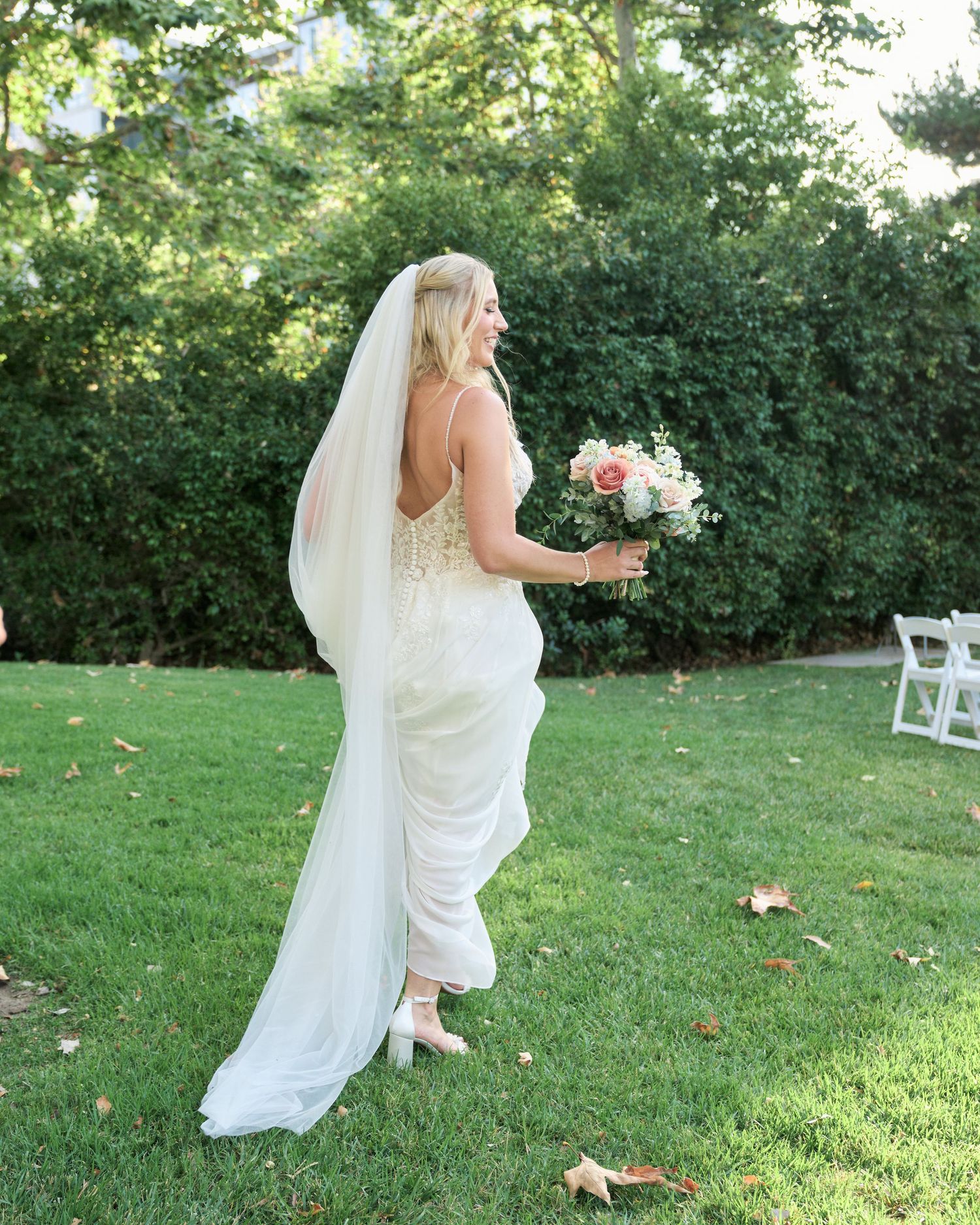 Bride in flowing white dress holds a bouquet while walking on grass.