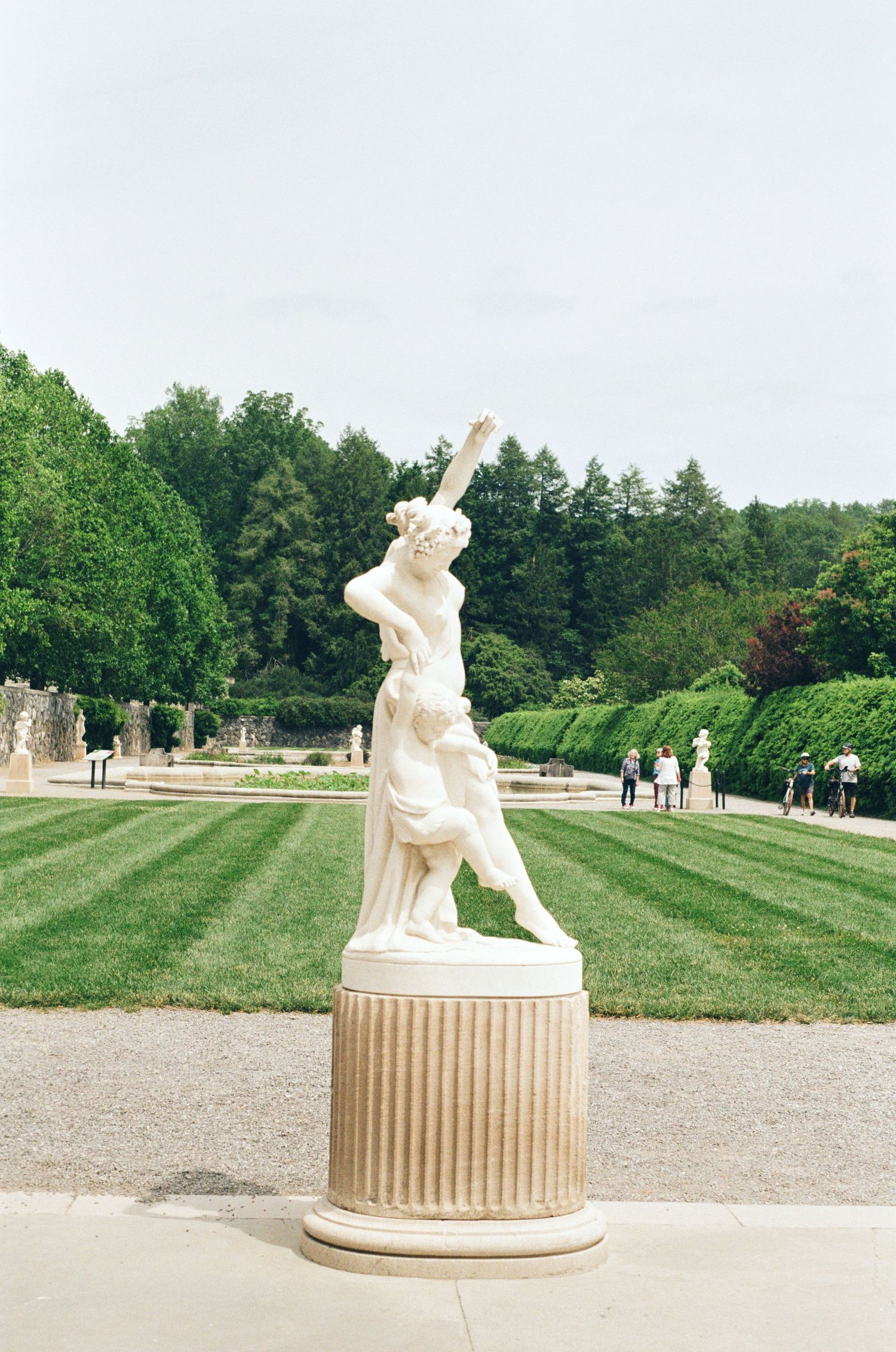 White marble statues on pedestals line a manicured garden path with green lawns and trees at a formal estate.