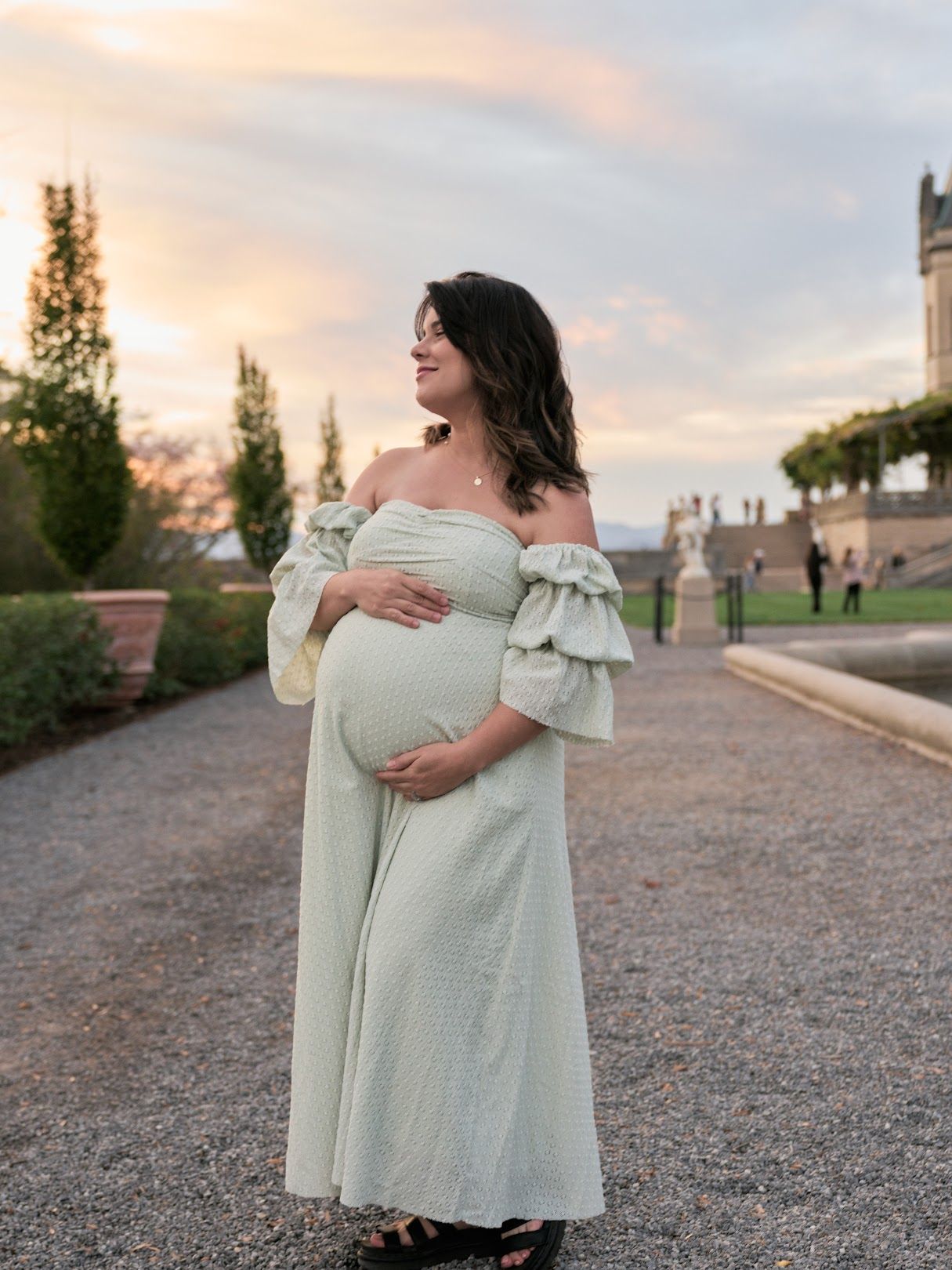 A pregnant woman in a white off-shoulder dress poses outdoors at sunset against a gravel path and trees.