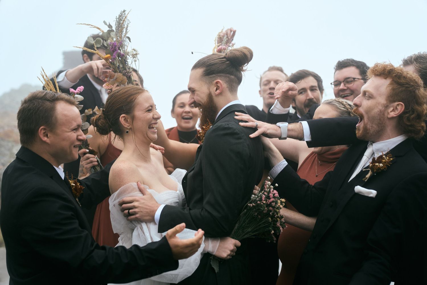A jubilant wedding party celebrates together outdoors with laughter and embraces.