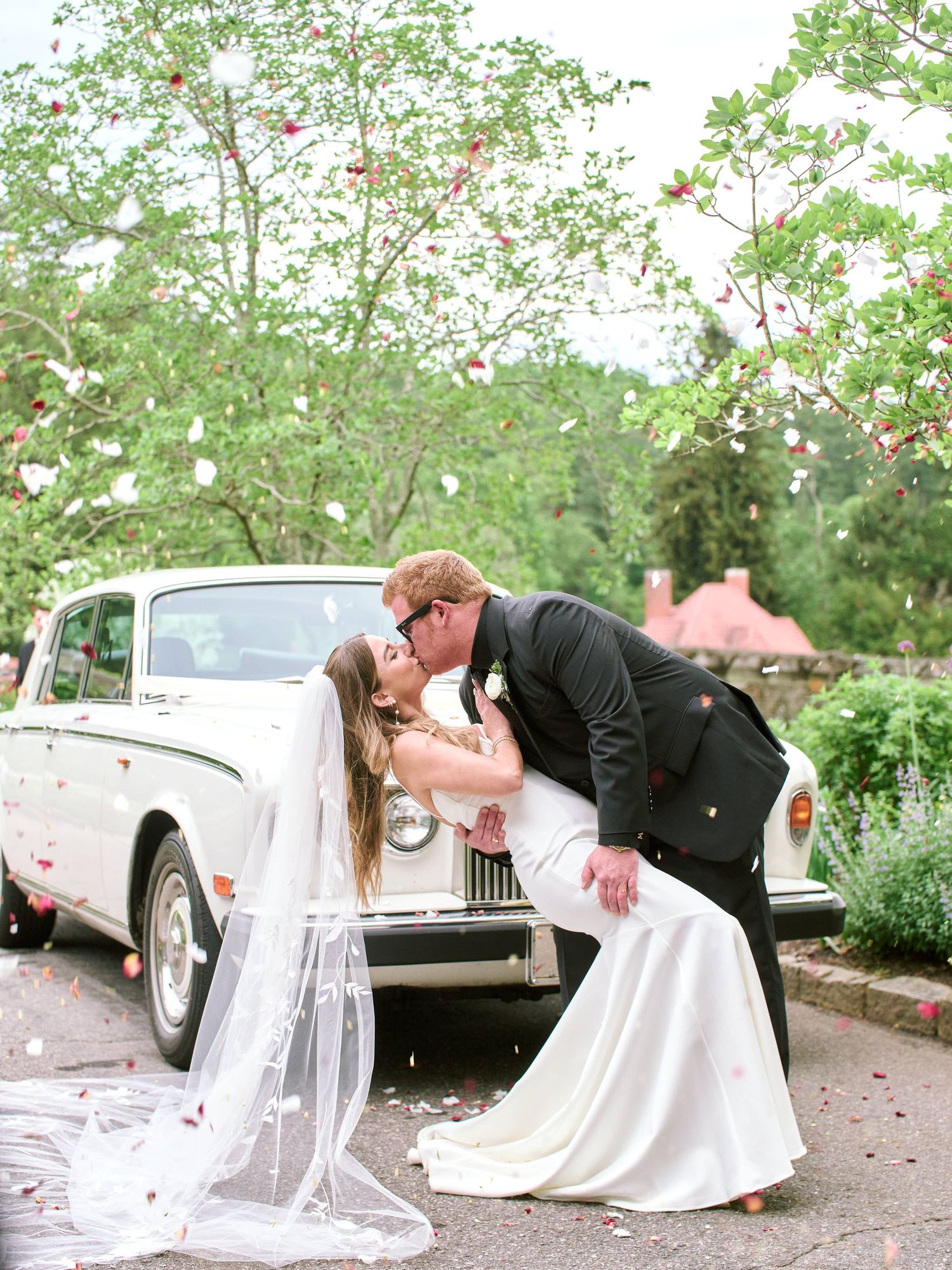 A romantic wedding couple shares intimate moments in a garden setting with vintage cars in the background.