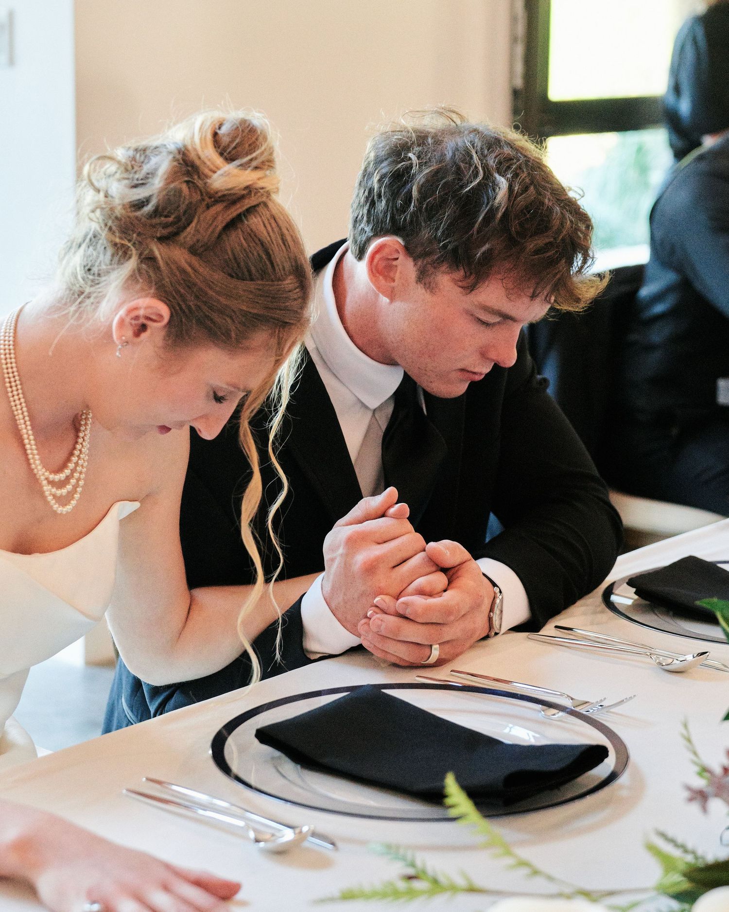 A couple bows their heads in prayer at an elegant dining table set with black charger plates and white linens.