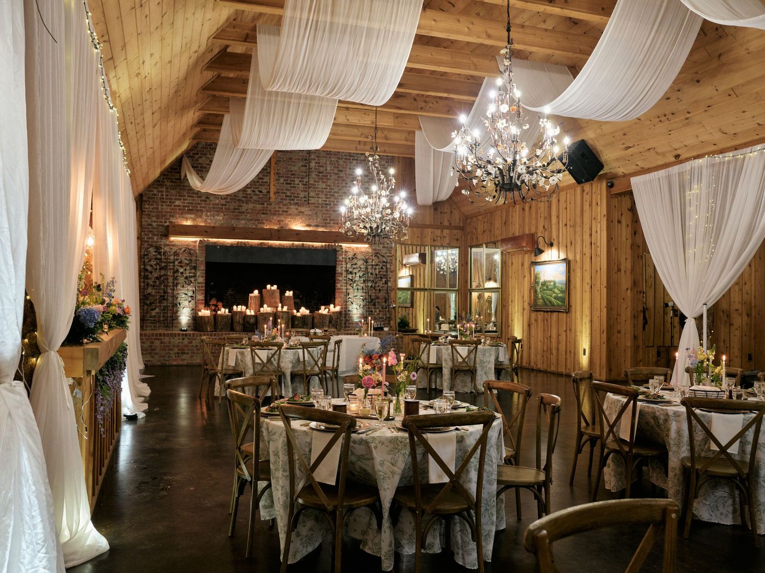 Rustic wedding reception venue with wood ceilings, draped white fabric, chandelier lighting, and elegantly set round tables.
