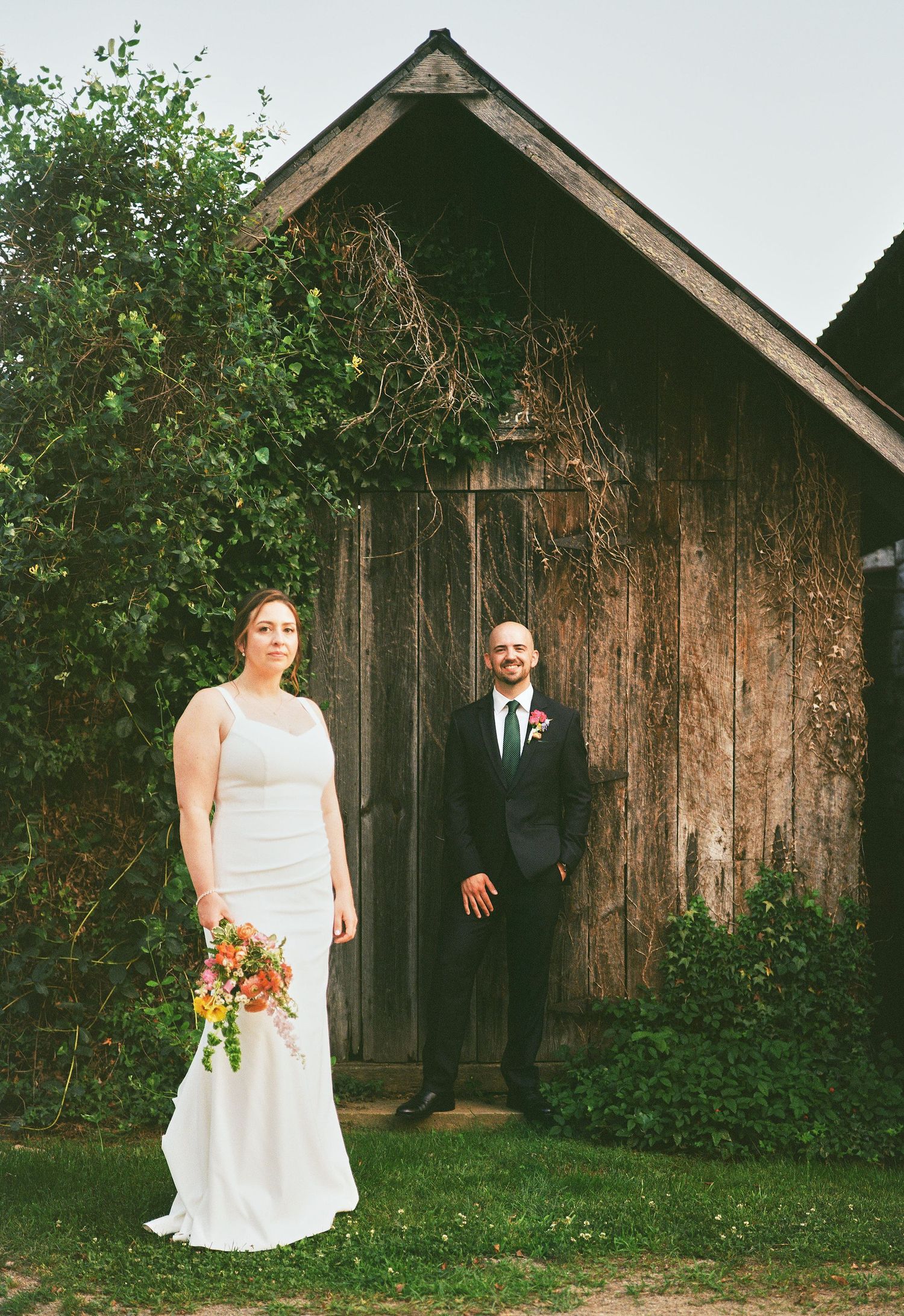 Bride in white gown holding colorful bouquet stands with groom in navy suit before rustic wooden barn.