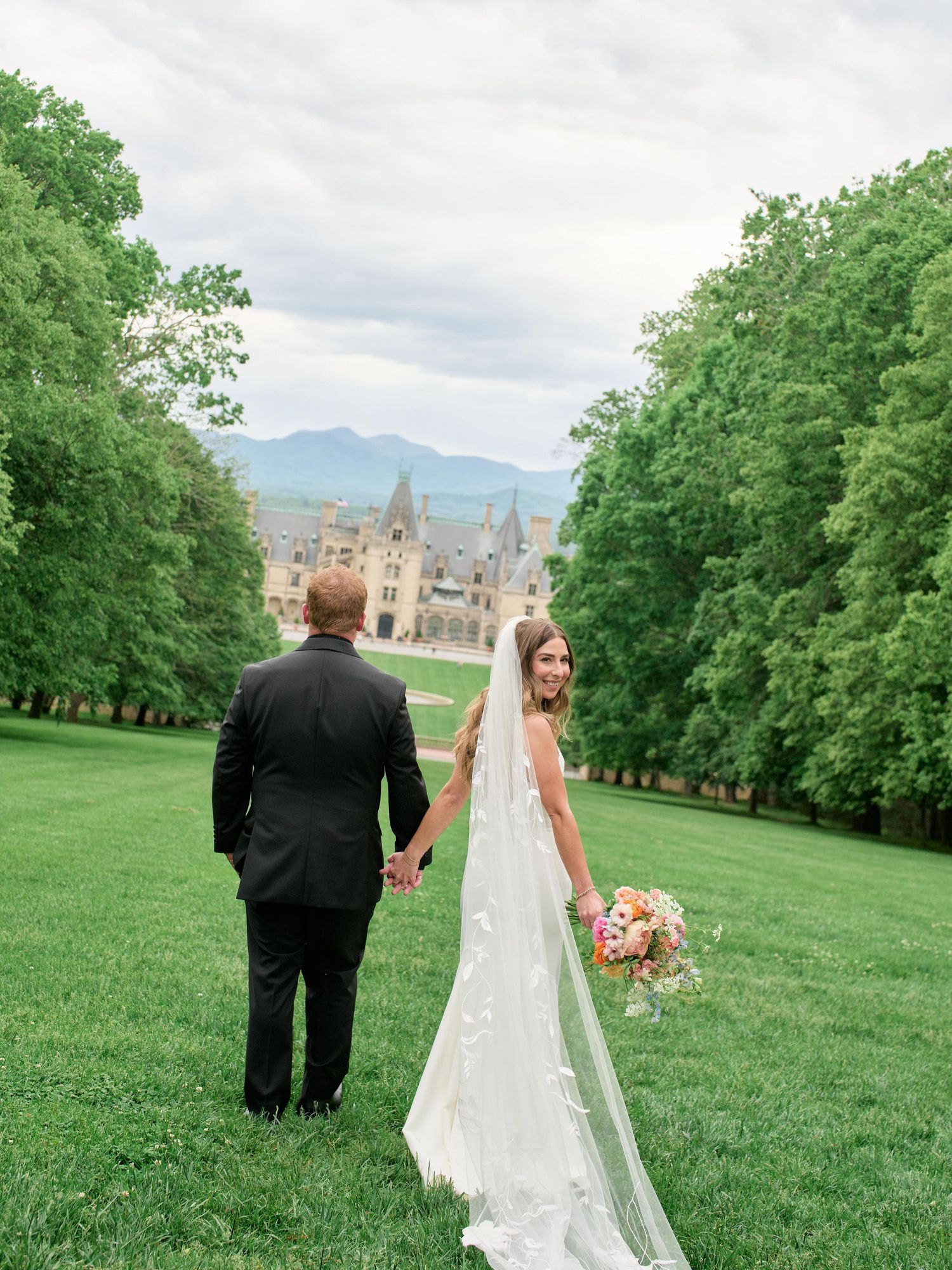 A couple in wedding attire walk on a lush green lawn with a grand castle-like building and mountains in the background.
