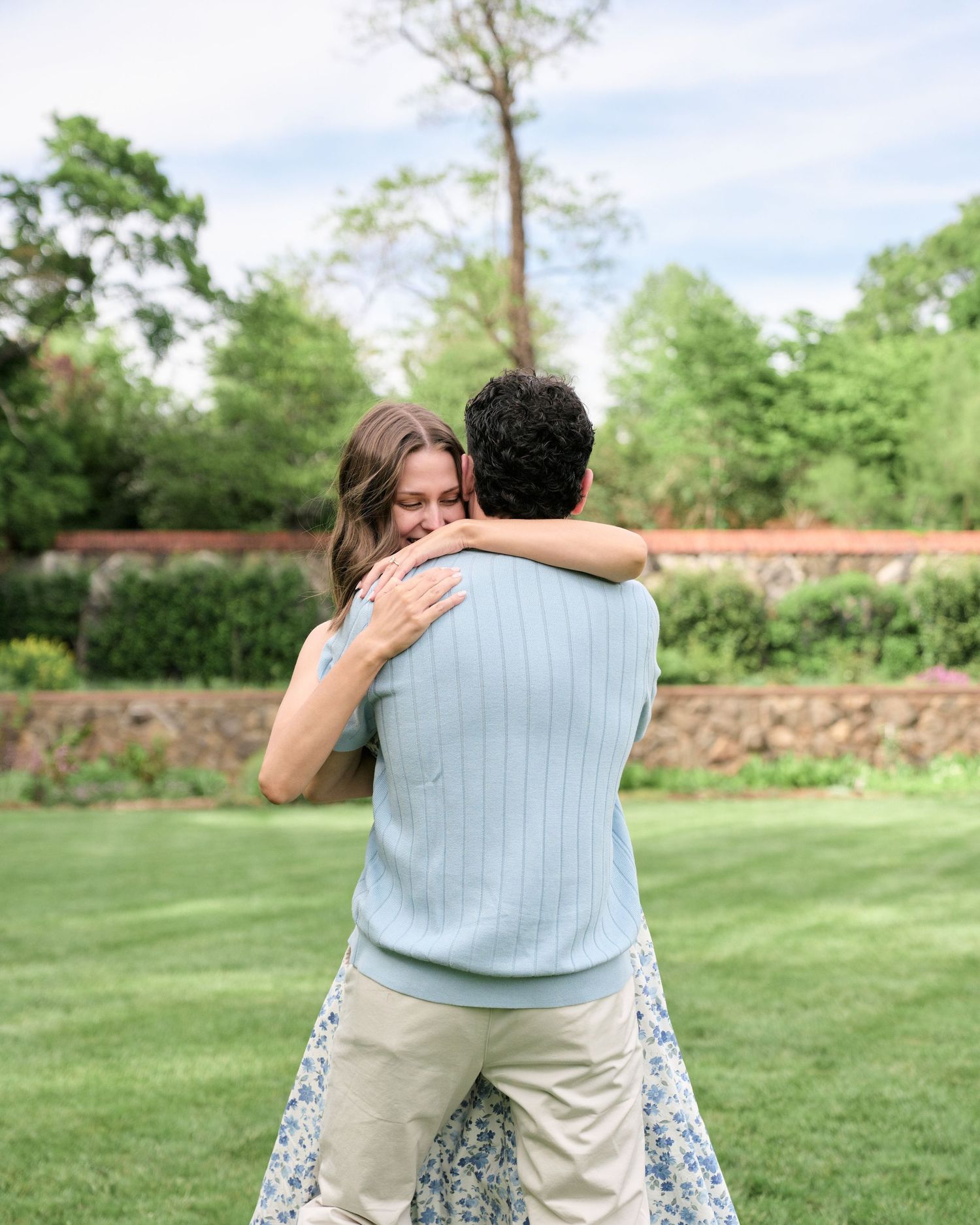 Couple embracing in a lush green garden, woman wearing floral skirt showing engagement ring, romantic outdoor proposal moment.