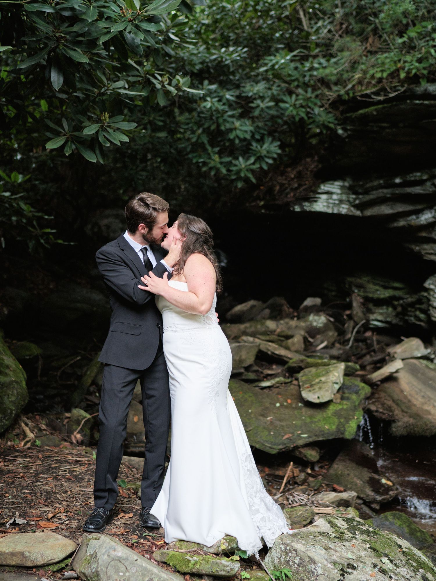 Bride and groom share a romantic kiss on mossy rocks near a waterfall surrounded by lush green trees.