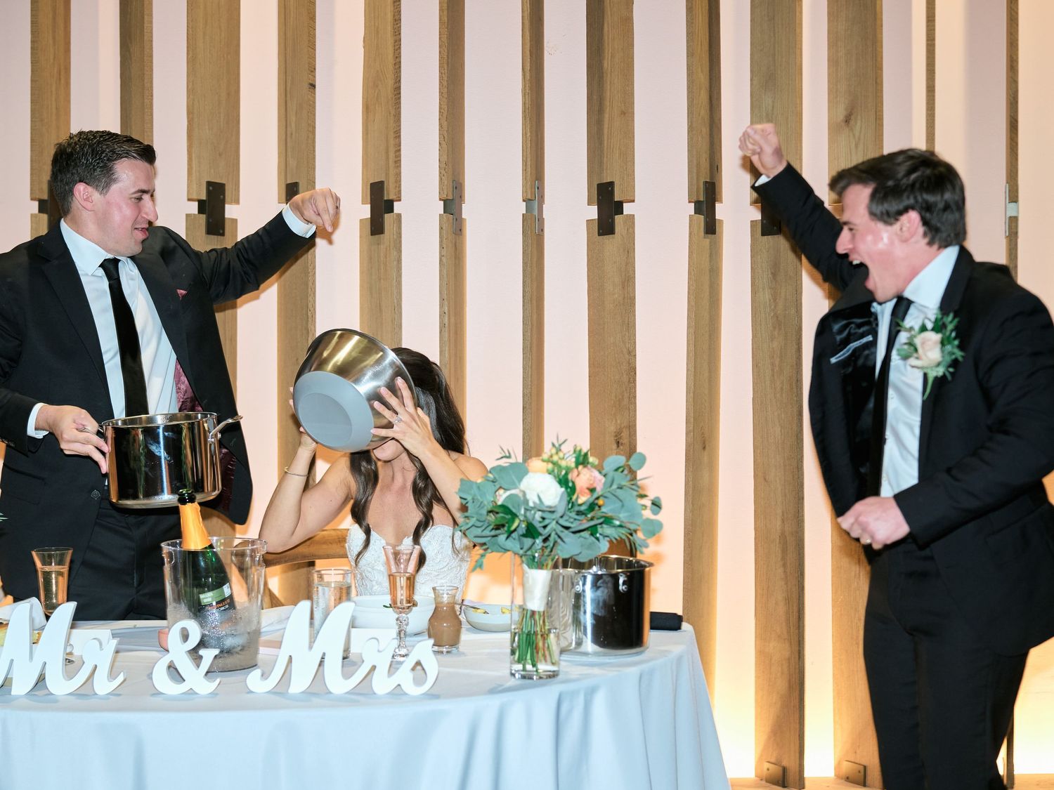 Joyful wedding reception celebration at Mr & Mrs table with guests cheering and bride covering her face.