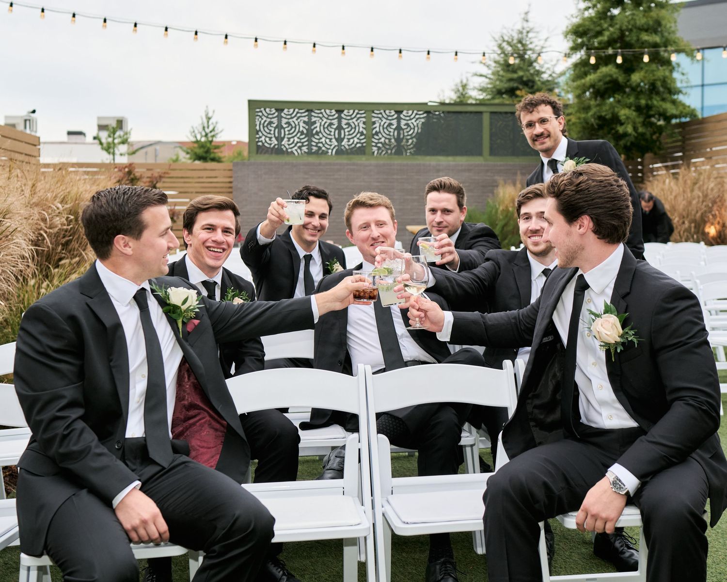 Groomsmen in black suits toasting with drinks at an outdoor wedding ceremony, seated on white chairs.