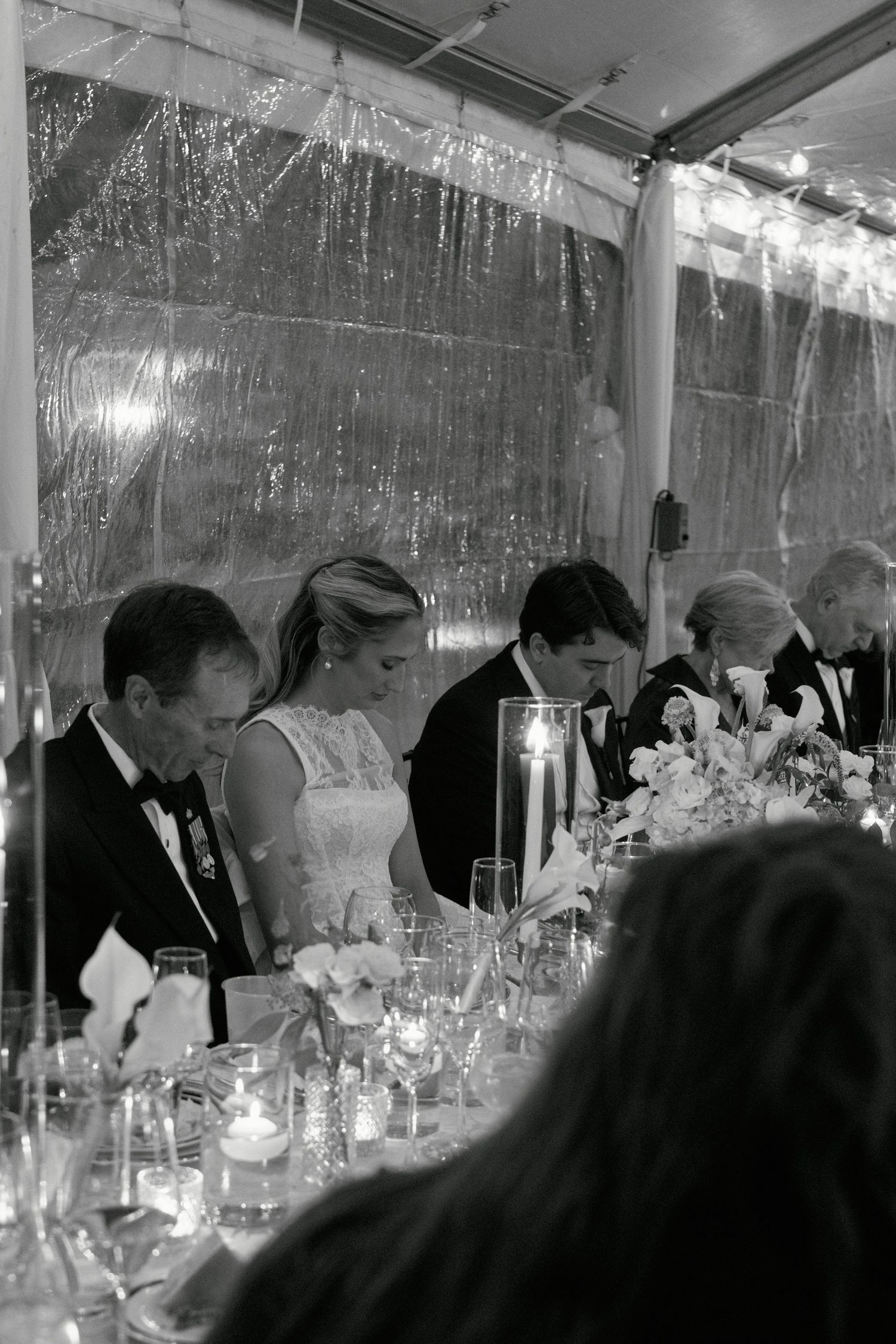 Black and white photo of bride and groom seated at wedding reception head table under a sparkling tent.