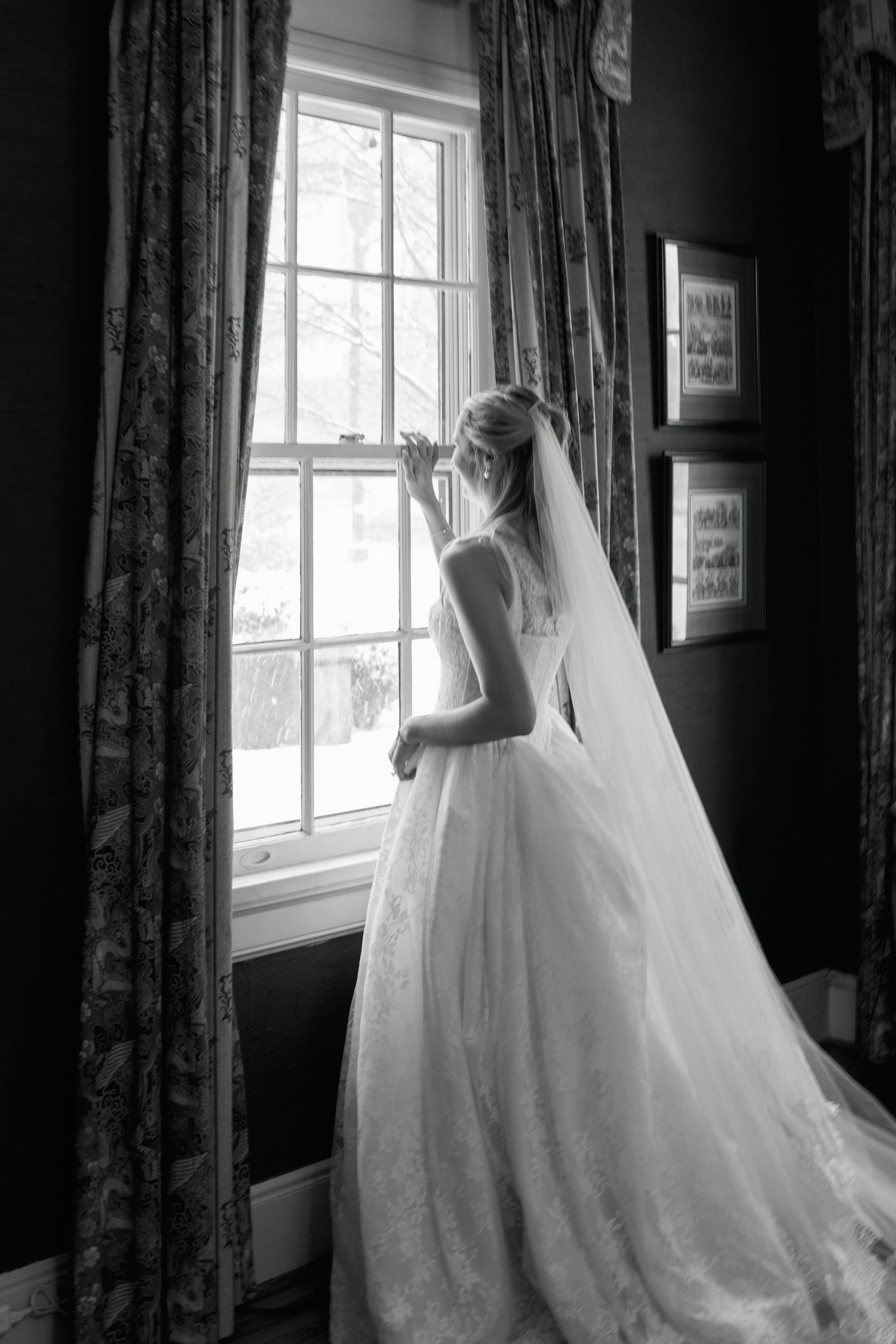 Black and white photo of a bride in a flowing gown and veil gazing out a window on her wedding day.