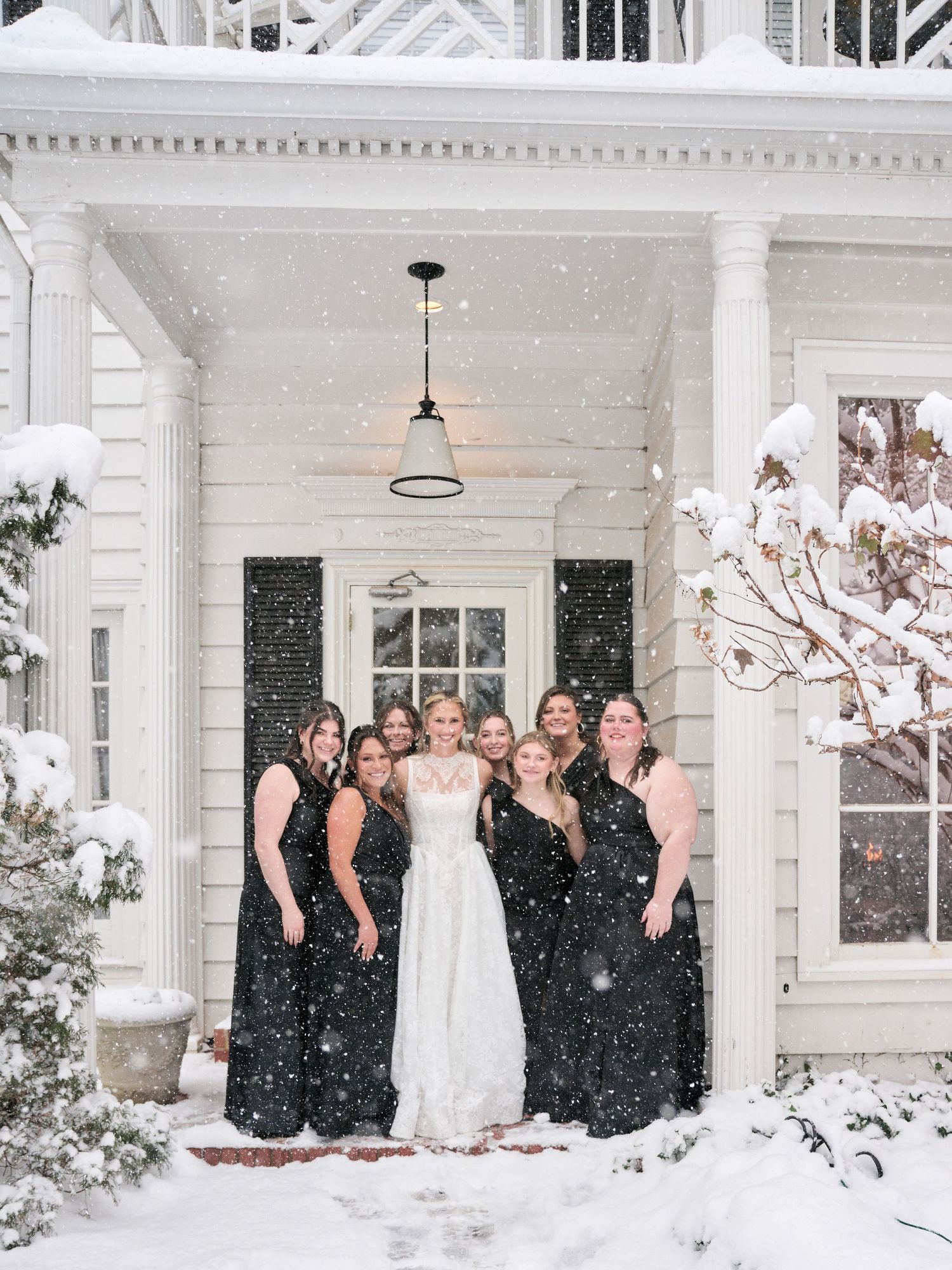 Bride in white gown with bridesmaids in black dresses posing on snowy porch during winter wedding.