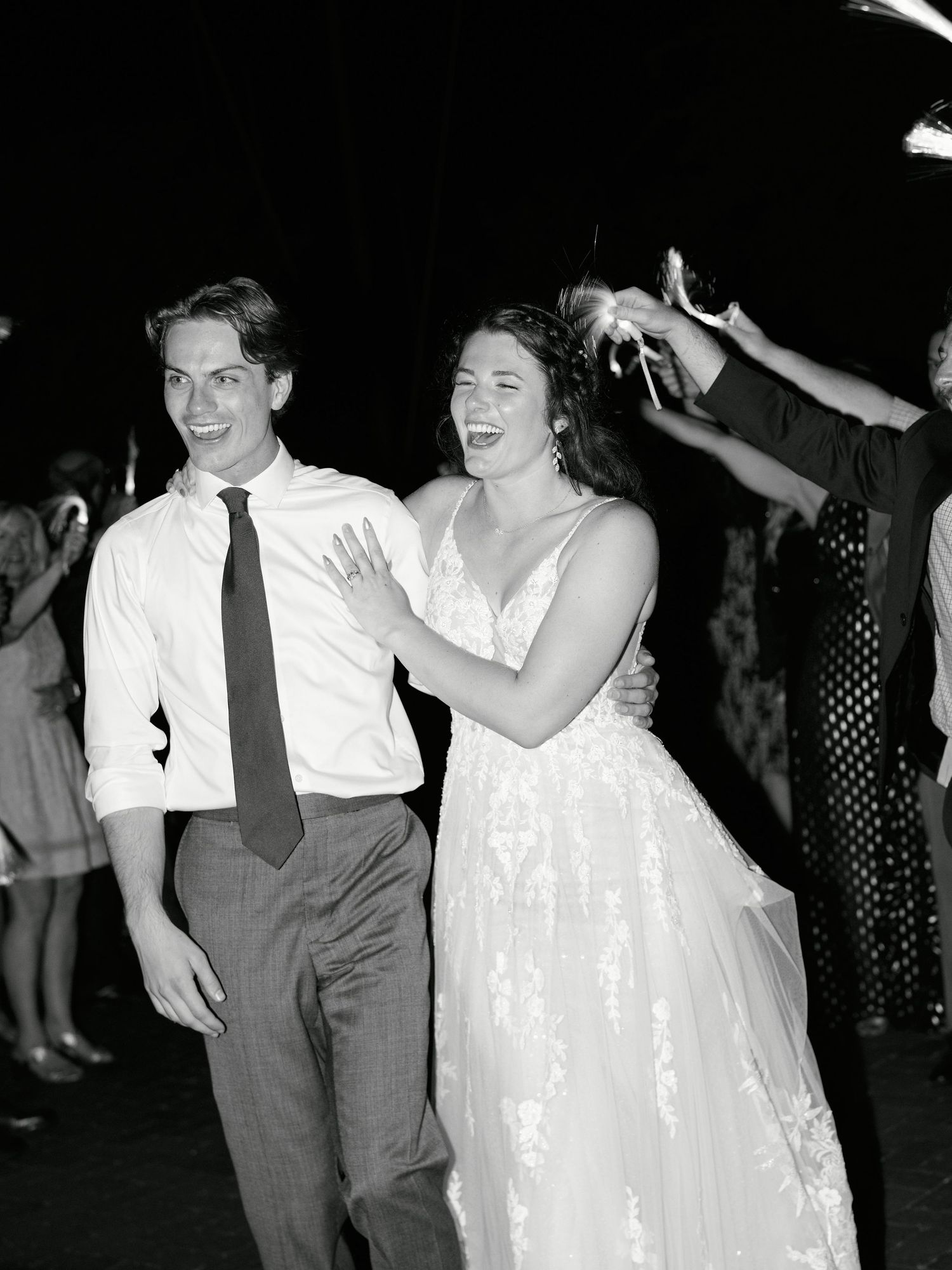 Black and white photo of a smiling bride in lace gown and groom in tie walking through sparkler exit at wedding reception.