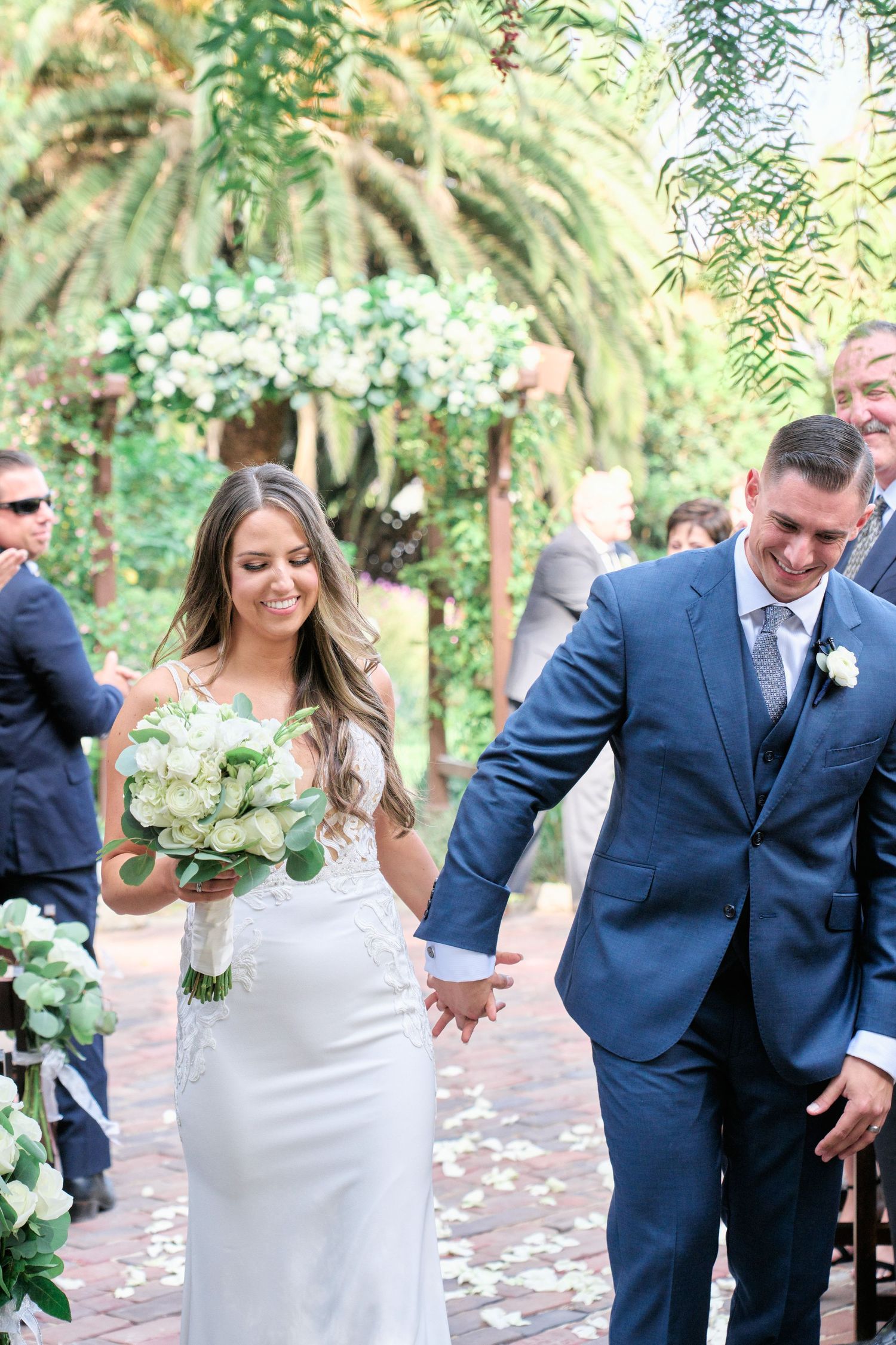 A couple walks hand in hand after their garden wedding ceremony surrounded by lush greenery and white flowers.