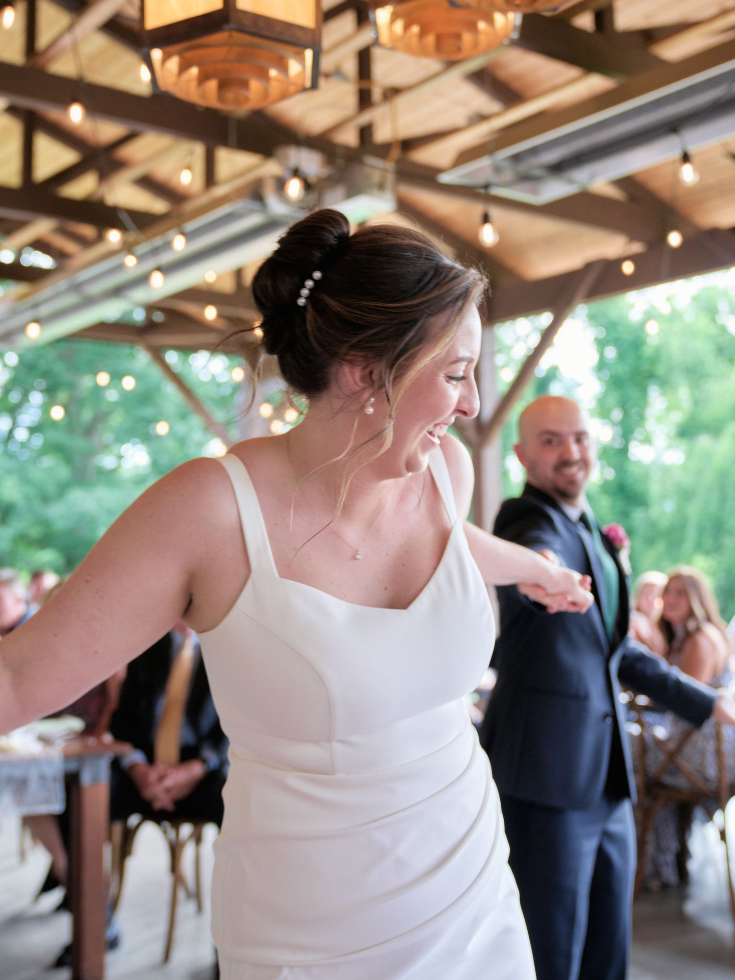Bride in elegant white dress dances joyfully under rustic pavilion with wooden beams.