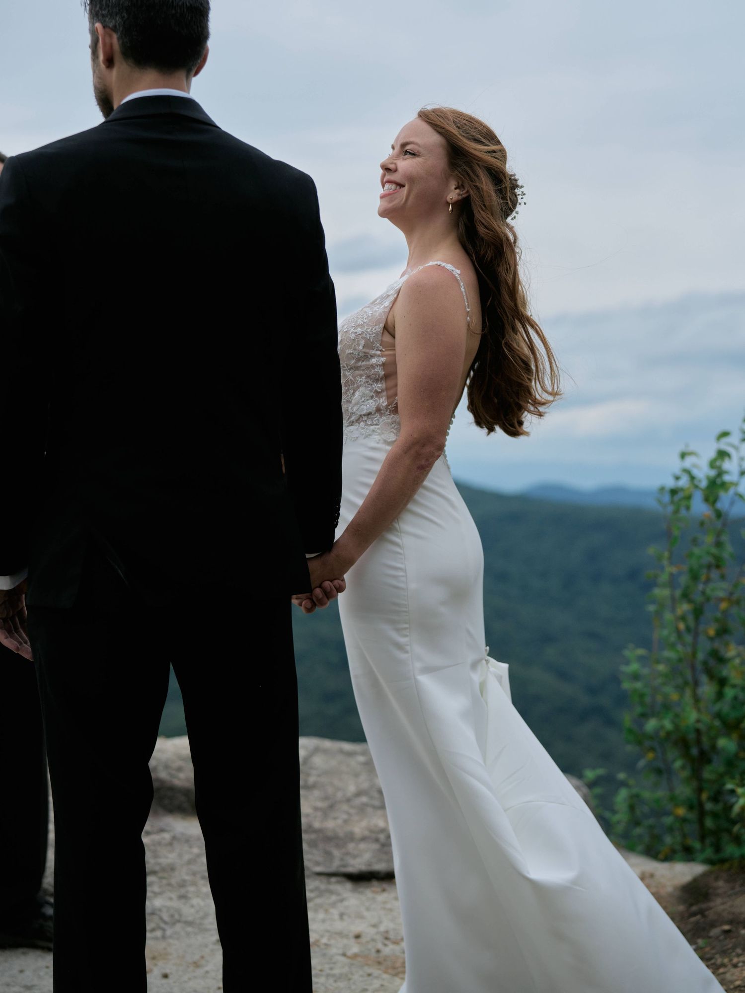A bride in a white dress laughs joyfully while holding hands with a groom in black on a mountaintop overlook.