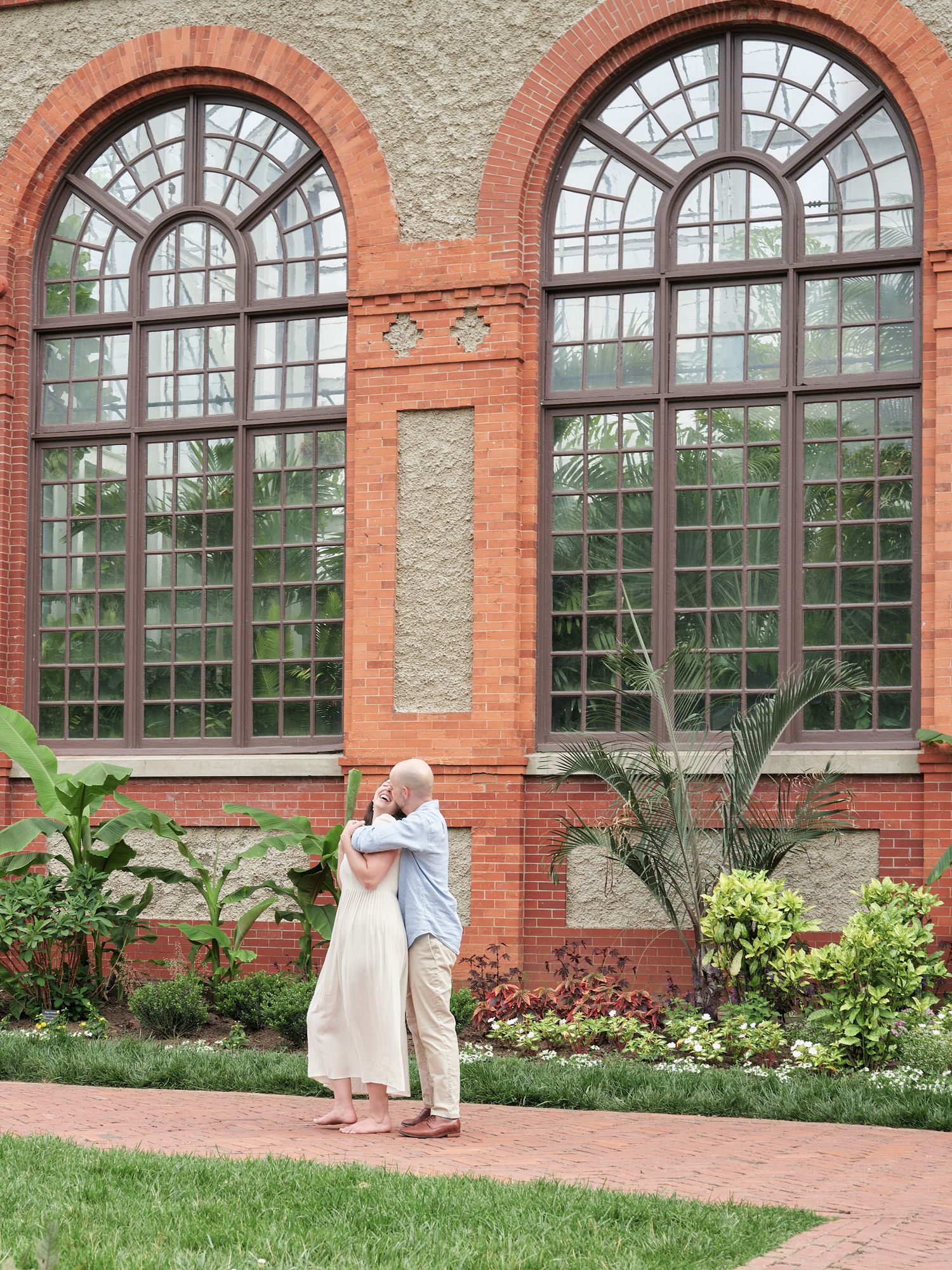 A couple embraces on a garden path in front of a historic building with arched windows.
