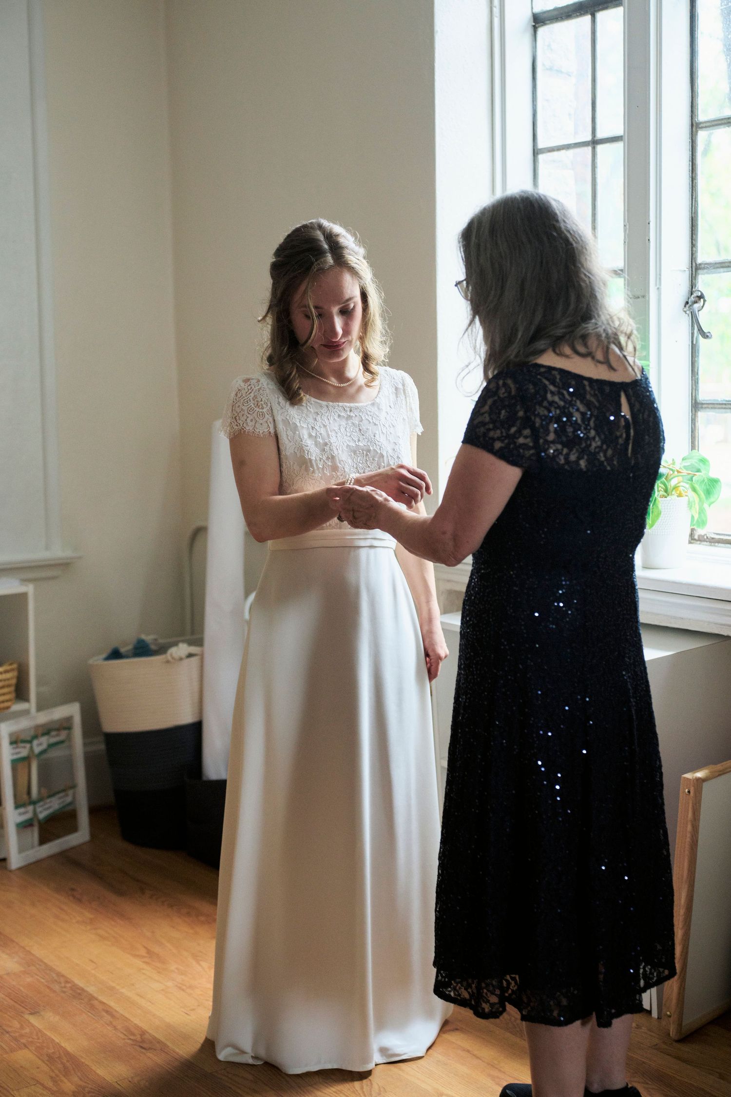 Two women stand near a window as one wearing a white wedding dress examines jewelry or rings with another in a black dress.