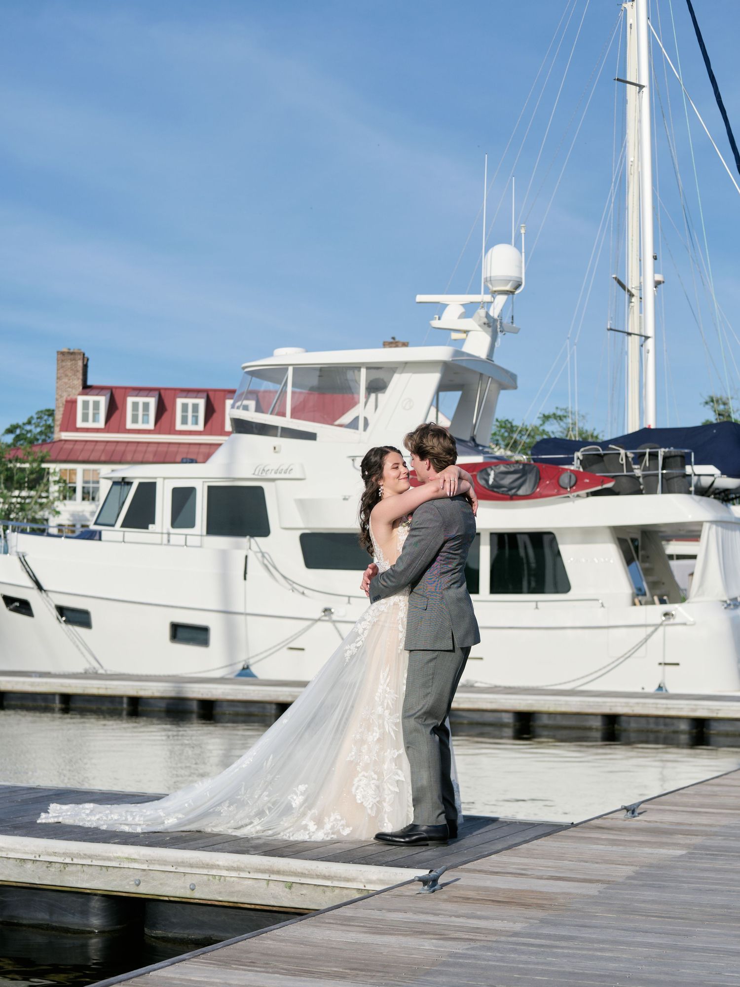 A couple embraces on a wooden marina dock with boats and a waterfront house in the background on a sunny day.