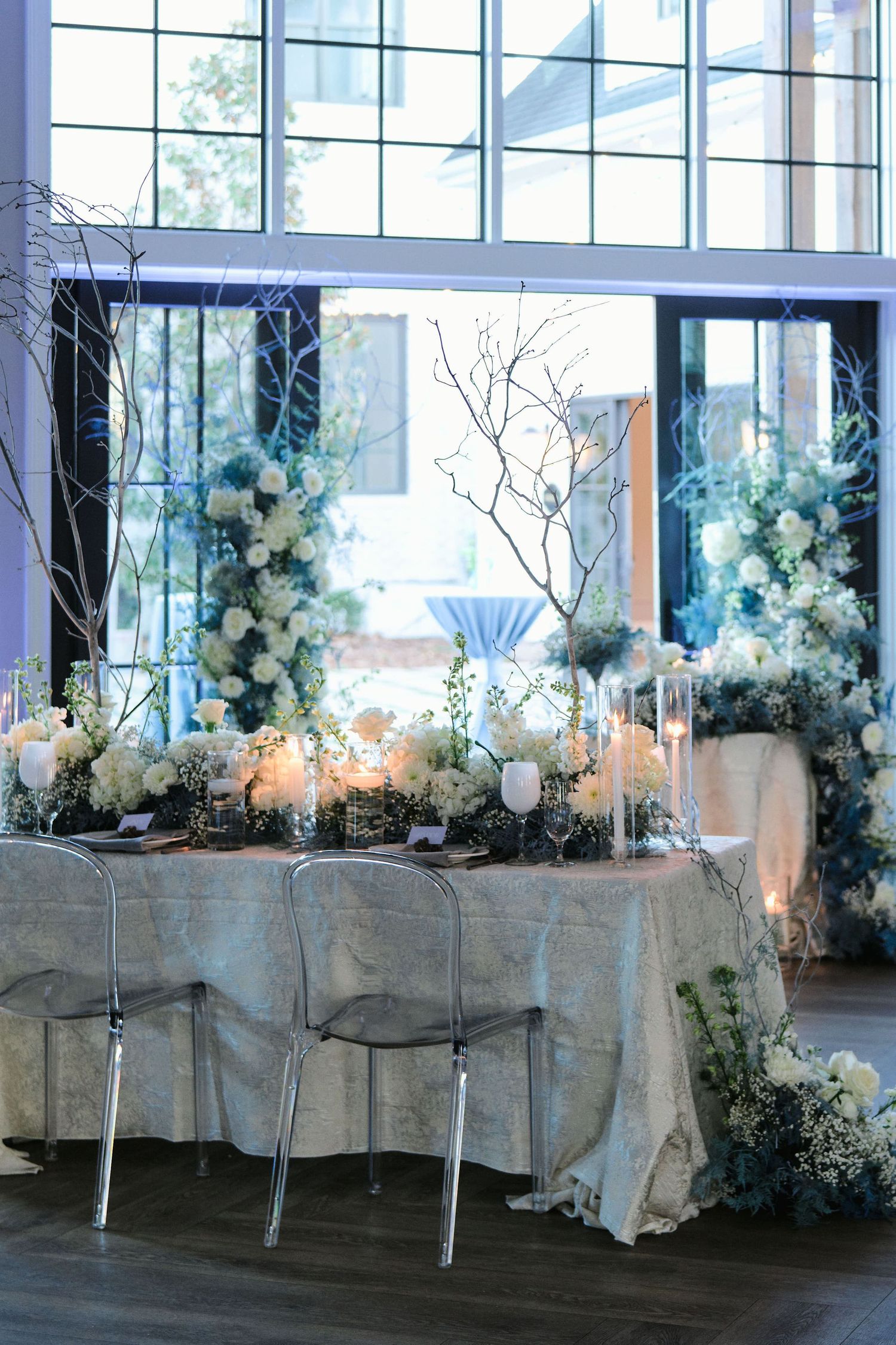Wedding table decorated with ethereal blue and white florals, branches, and ghost chairs.
