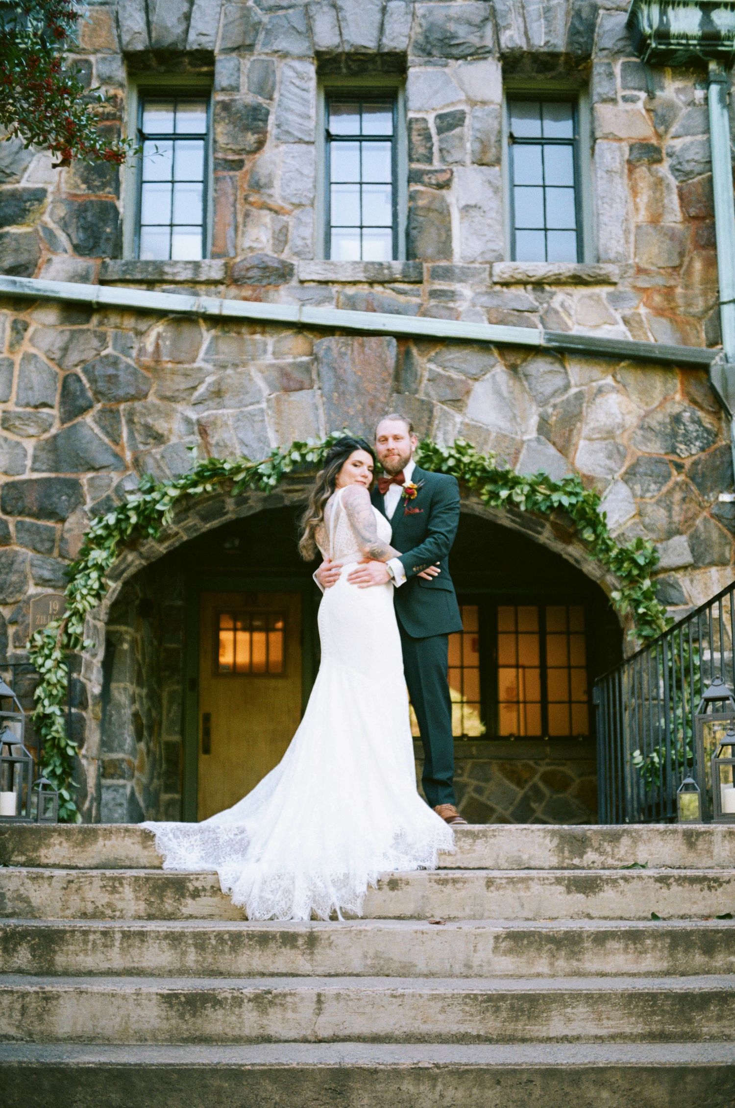 Wedding couple poses on stone staircase in front of elegant castle-style building with arched entrance.