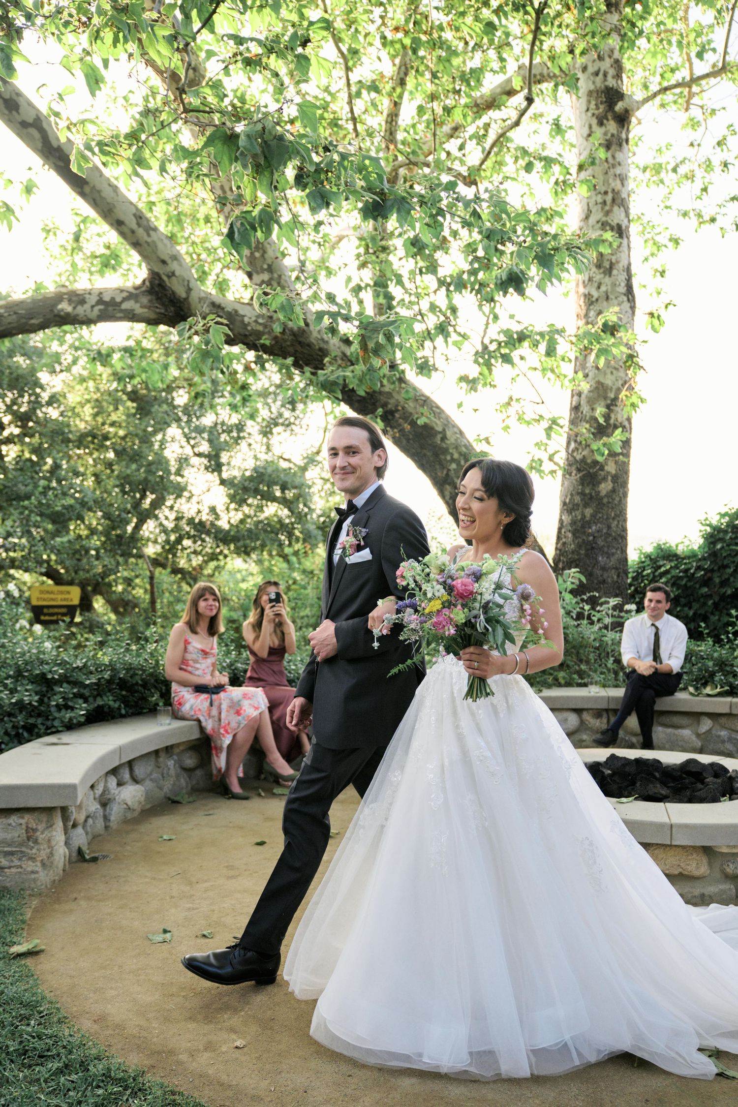 A wedding couple makes their entrance into a garden ceremony space with guests seated nearby.