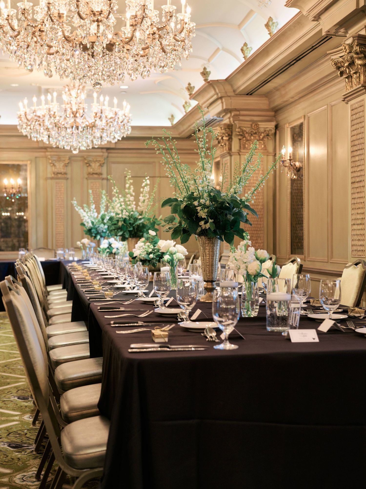 Luxurious wedding reception table setting with crystal chandeliers and tall floral centerpieces.