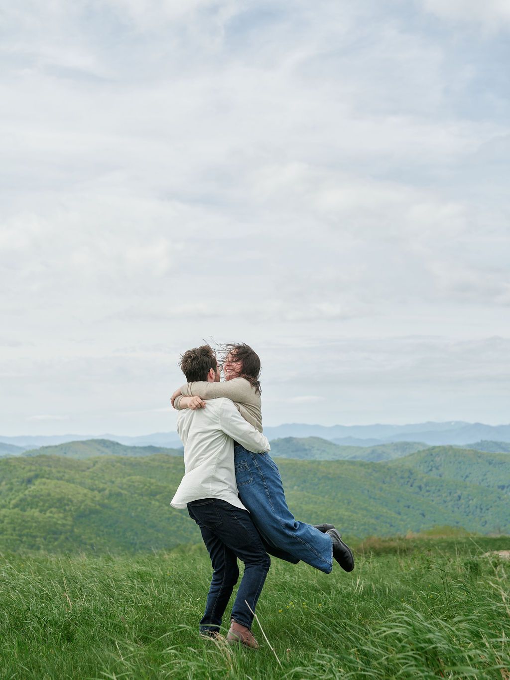 A loving embrace on a hilltop overlooking rolling green mountains under a cloudy sky.