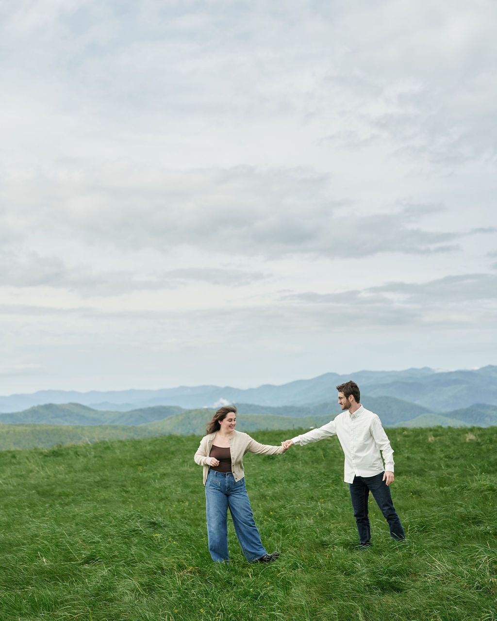 Two people dance and spin together on a grassy hilltop against a backdrop of misty blue mountains on an overcast day.