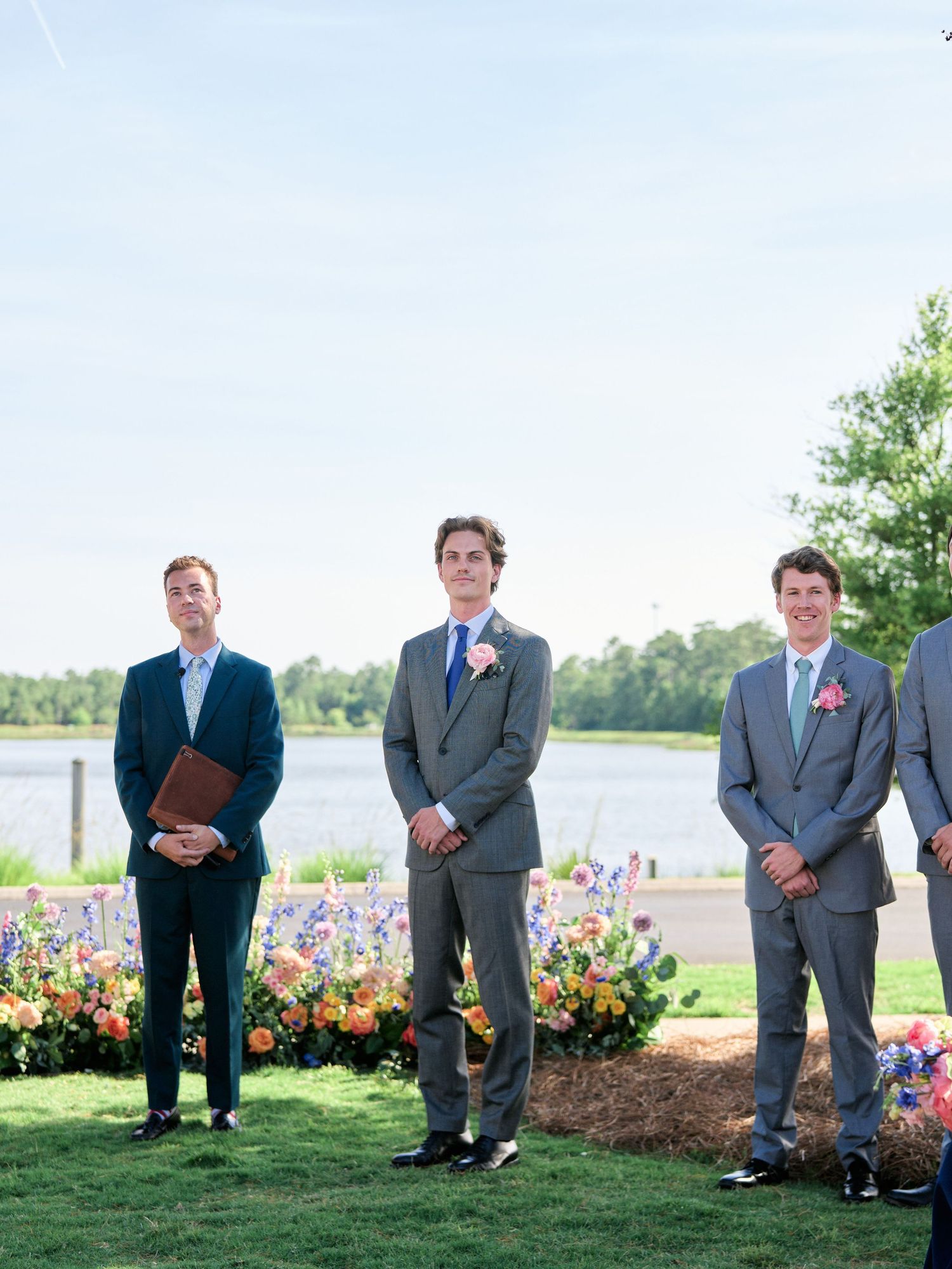 Three men in suits stand outdoors near a lake with colorful flowers in the foreground on a sunny day.