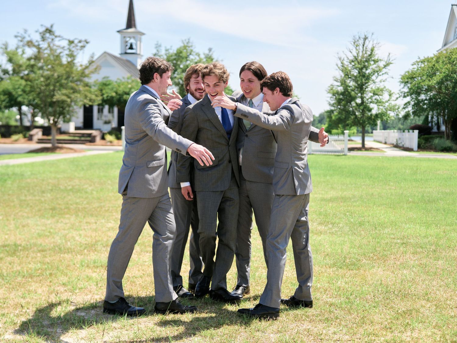 Group of groomsmen in grey suits sharing a moment on grass lawn.