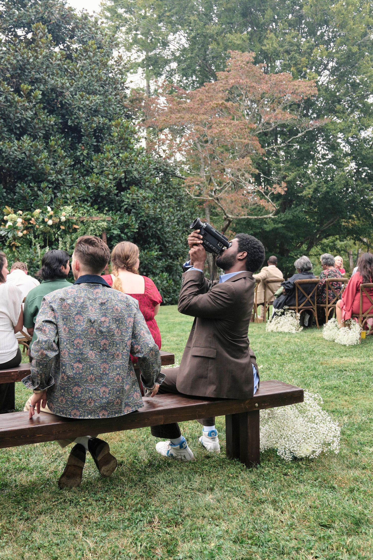 Guests enjoying an outdoor wedding reception seated at wooden benches under tree canopy.
