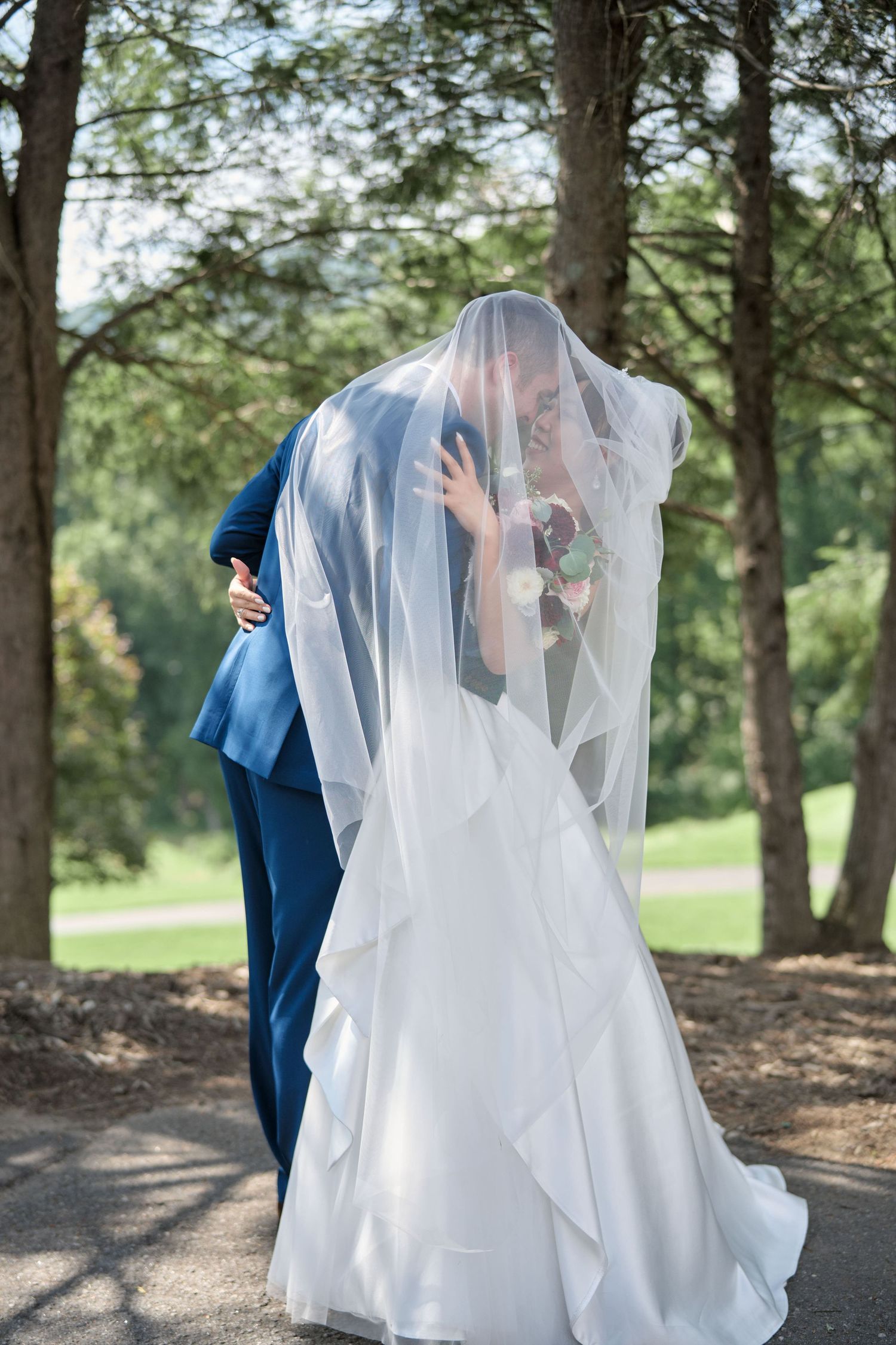 Romantic outdoor wedding photo with flowing veil and navy suit against a forest backdrop.