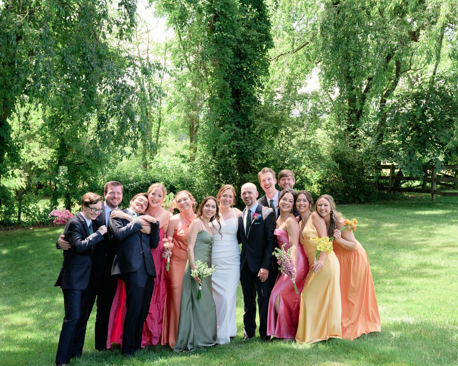 Wedding party poses together on lush green lawn wearing colorful formal attire in vibrant outdoor setting.