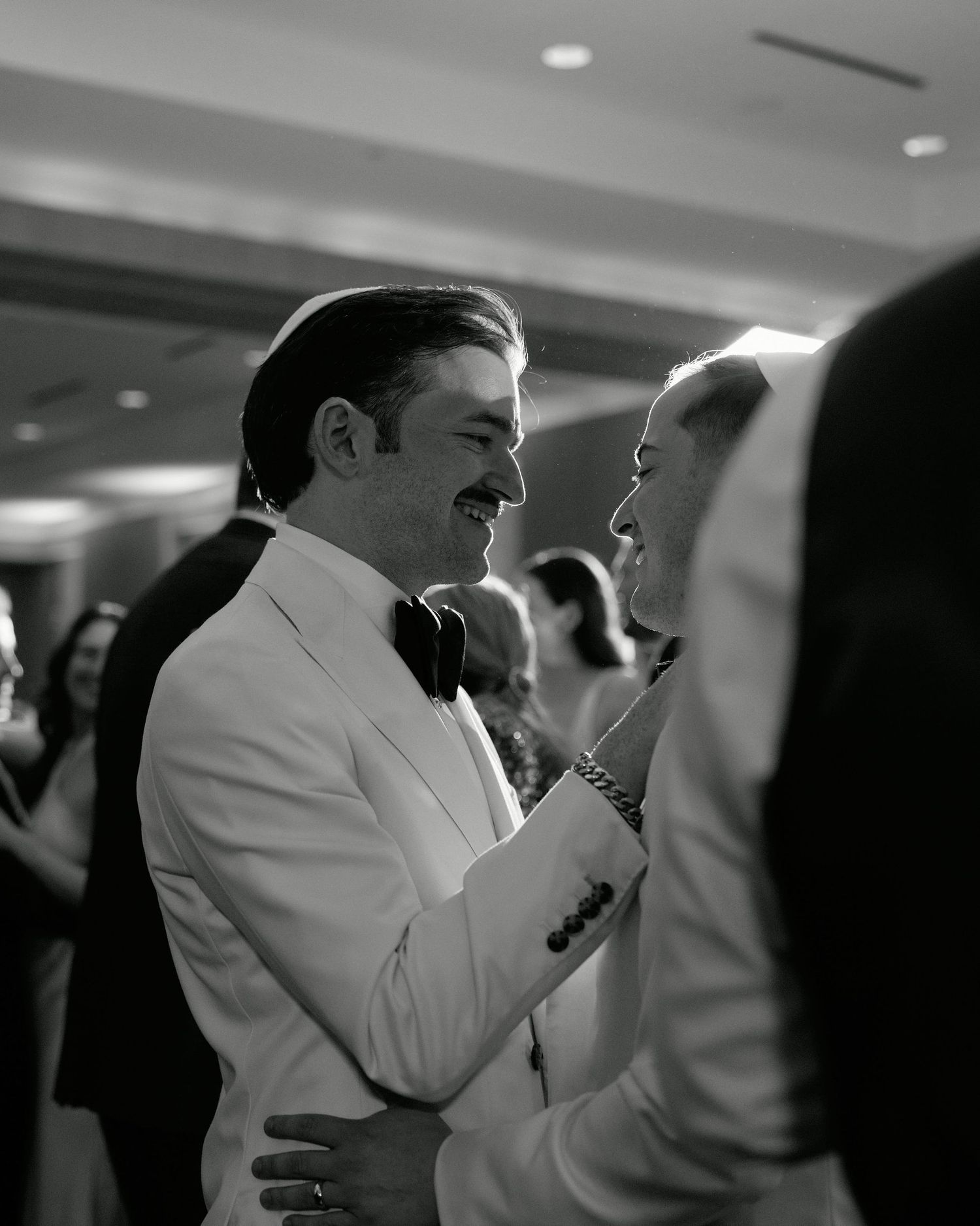 A joyful black and white candid moment between a man in a white tuxedo jacket and a wedding guest at a reception.
