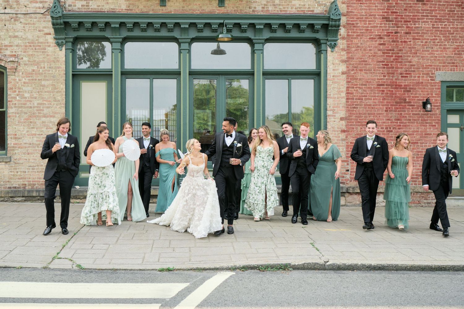 A wedding party poses together in formal attire against a brick wall with green trim and large windows.