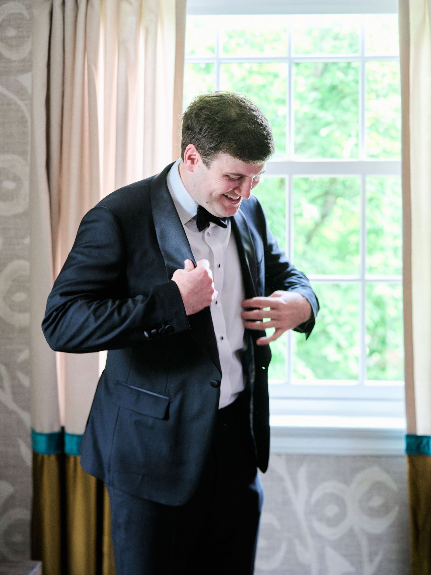 A person in a black tuxedo adjusts their formal attire by a bright window.