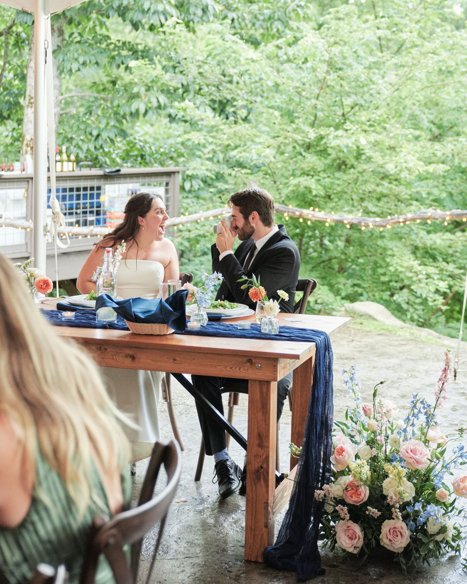 Bride and groom seated at rustic wooden sweetheart table with navy blue runner and floral arrangements at outdoor wedding reception.