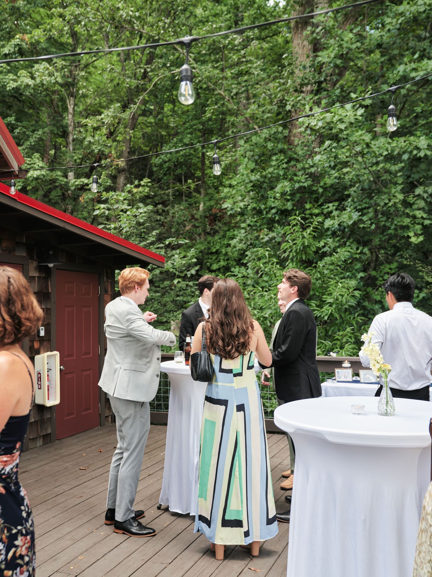 Guests mingle on an outdoor deck at a wedding cocktail hour, surrounded by lush green trees and string lights.