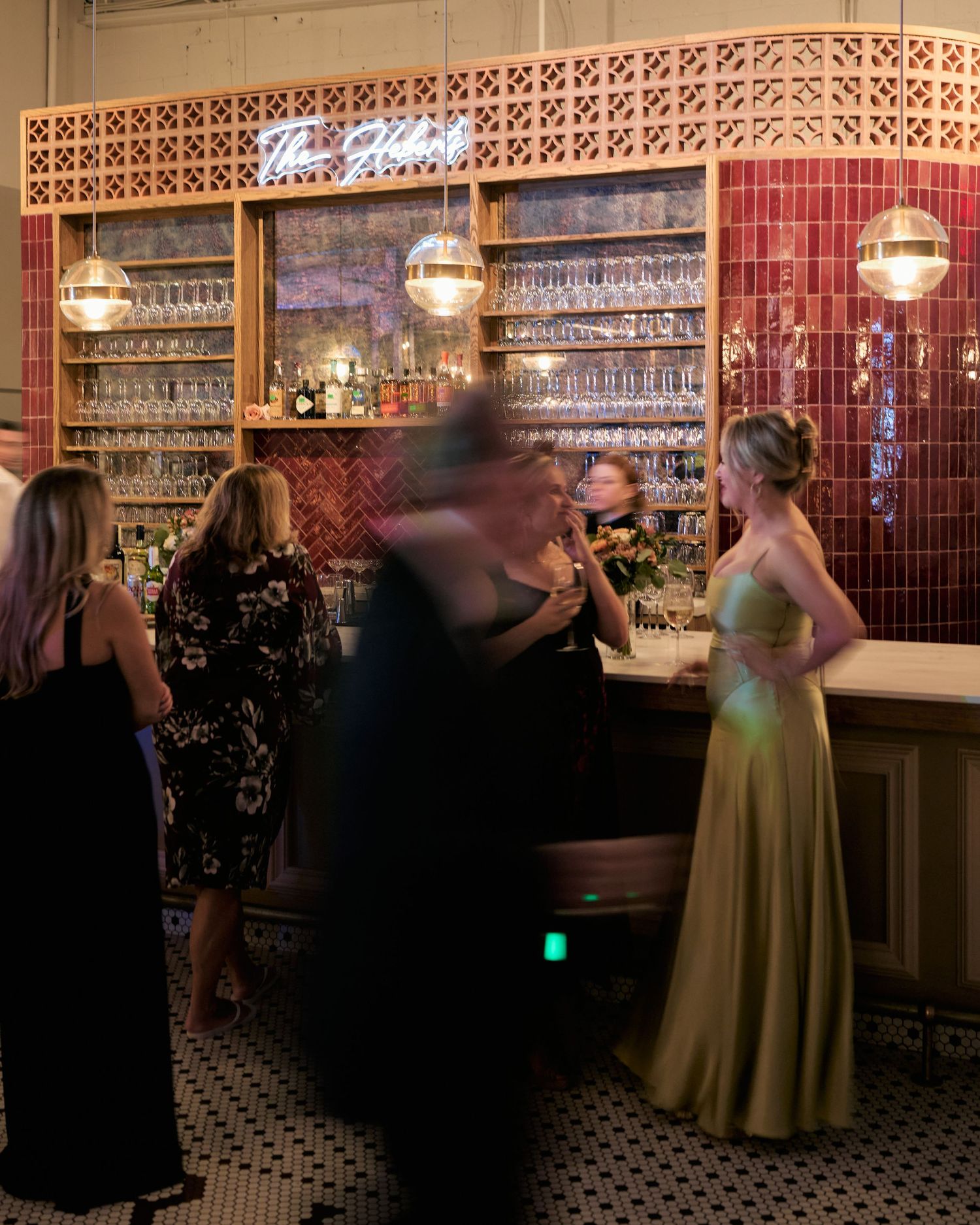 Elegant guests in formal attire gather at a stylish red-tiled bar with pendant lights and stocked shelves.