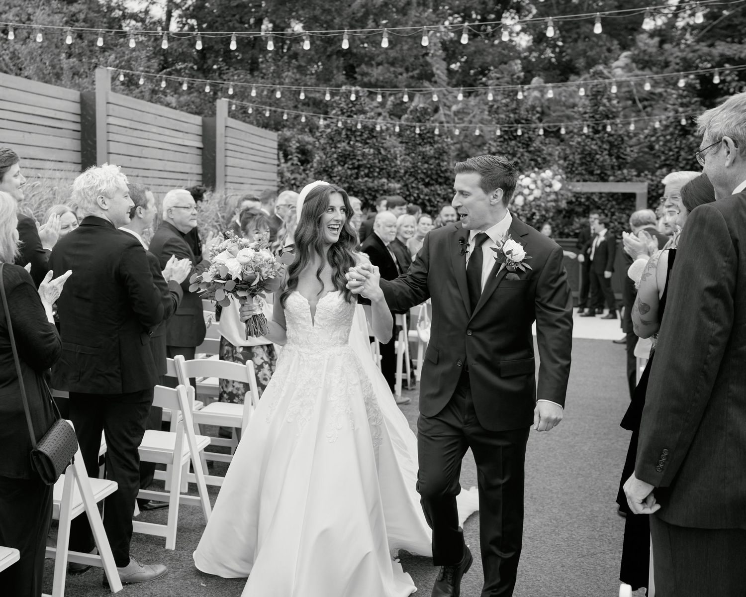 Bride and groom walk down the aisle at outdoor wedding ceremony with string lights and applauding guests.