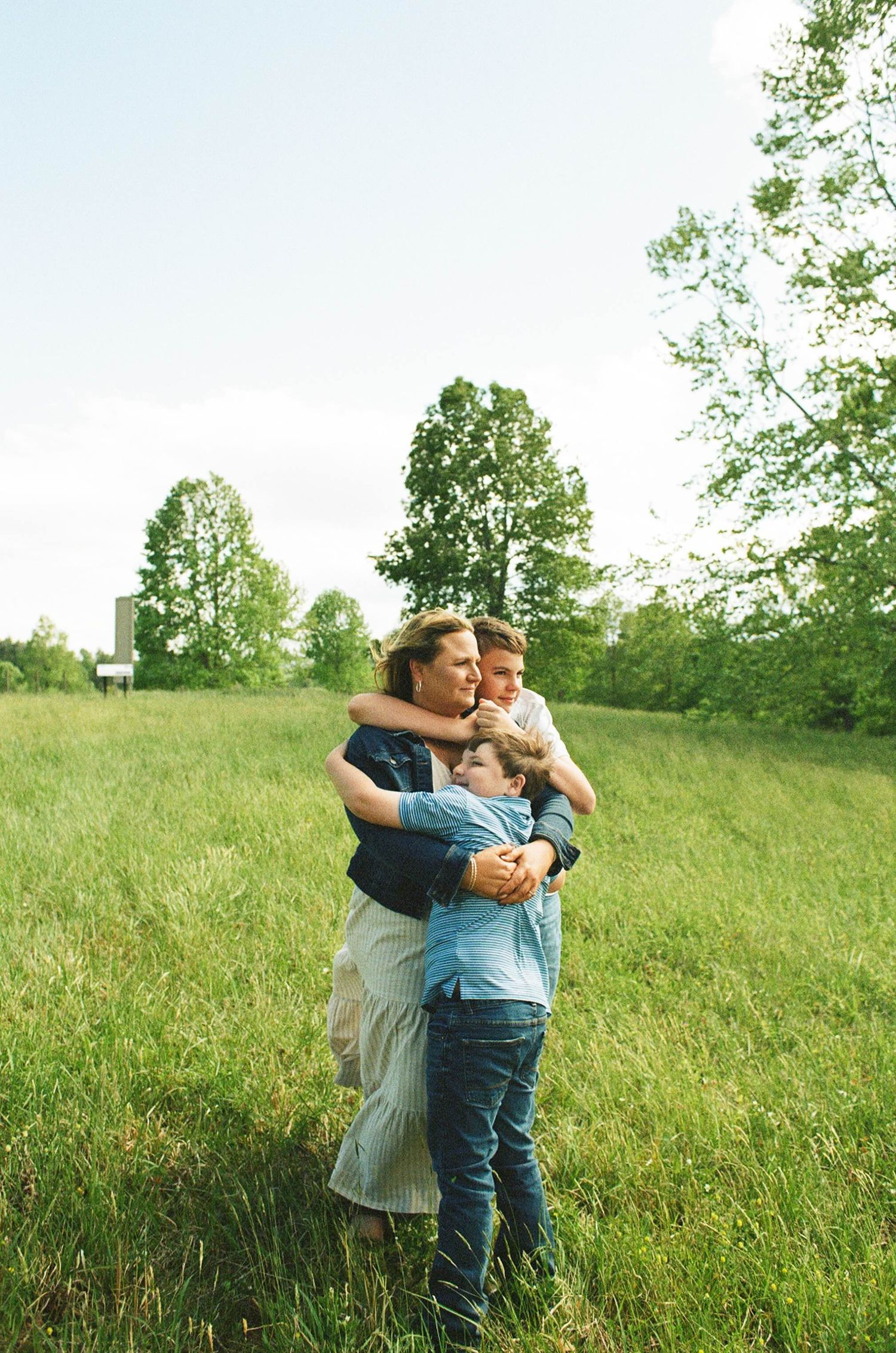 A family of three embraces in a lush green field surrounded by trees on a bright sunny day.