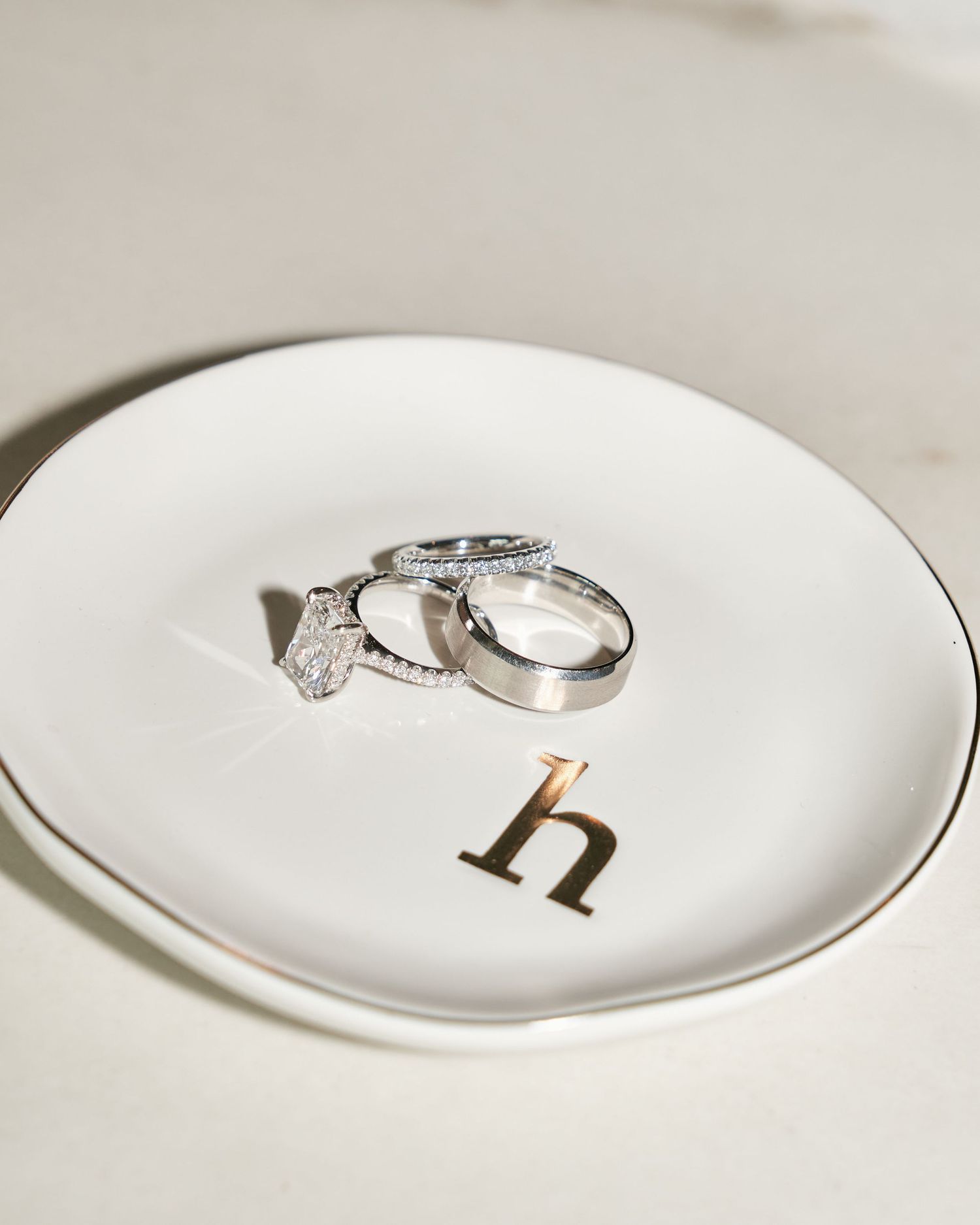 Three silver wedding rings and a diamond engagement ring rest on a white ceramic ring dish with a gold letter h.
