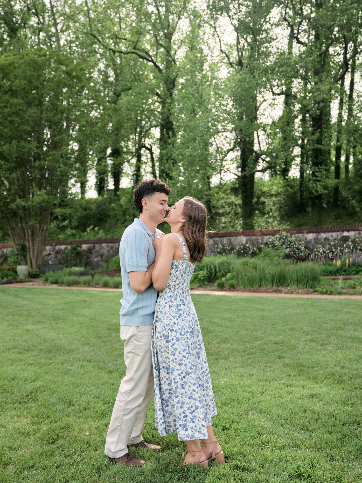 Young couple sharing romantic moment on garden lawn, woman in blue floral dress, surrounded by greenery.